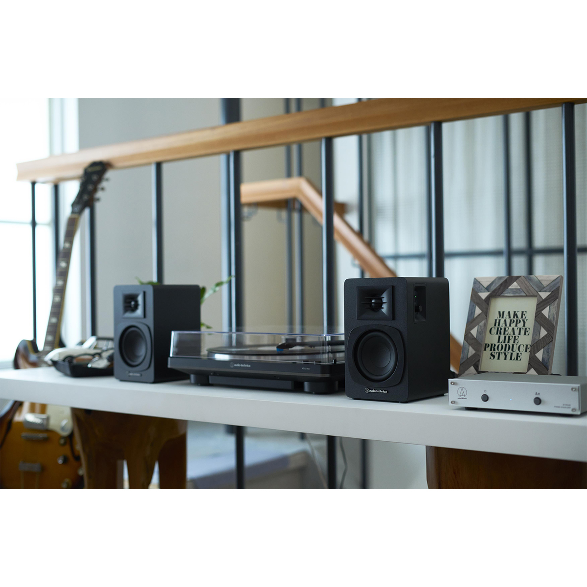 Biała półka z czarnymi głośnikami, gramofonem, gitarą i oprawionym cytatem. W tle drewniana balustrada.