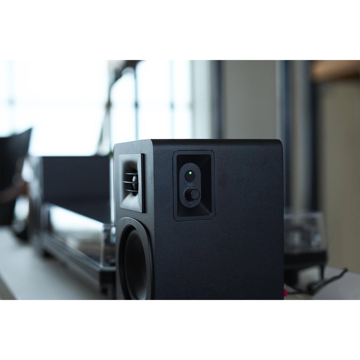 Czarny głośnik z regulacją głośności i zielonym światłem, umieszczony na białej powierzchni. W tle widoczny jest inny sprzęt audio.