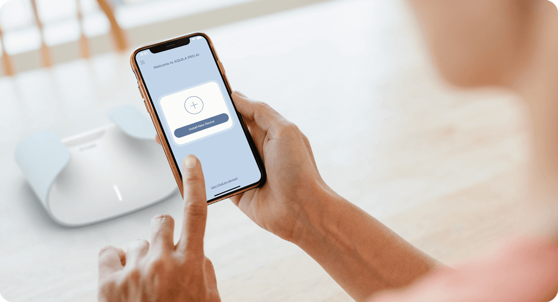 Osoba trzyma telefon z interfejsem aplikacji z ikoną plus i 'Zainstaluj nowe urządzenie'. Białe urządzenie leży na stole.