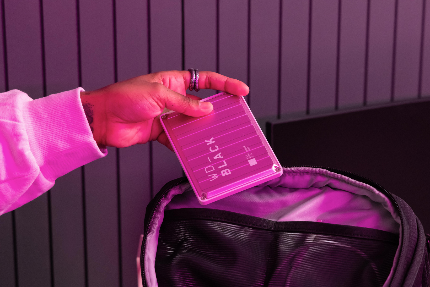 Una mano sostiene un disco duro externo WD_BLACK rosa, colocándolo en una mochila negra. La escena está iluminada con luz rosa.