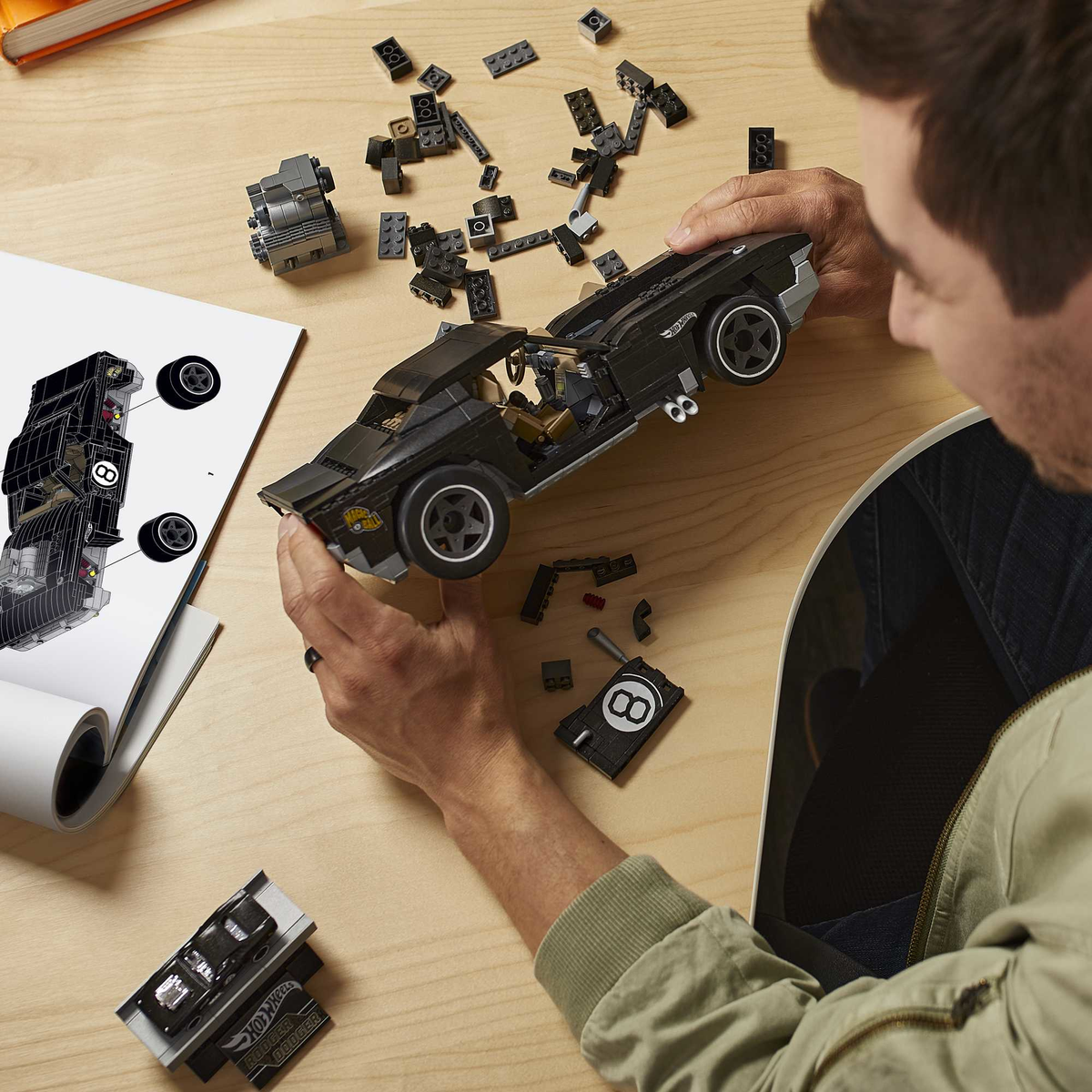 Osoba składająca czarne auto Lego na drewnianej powierzchni. Instrukcje i klocki Lego rozrzucone.