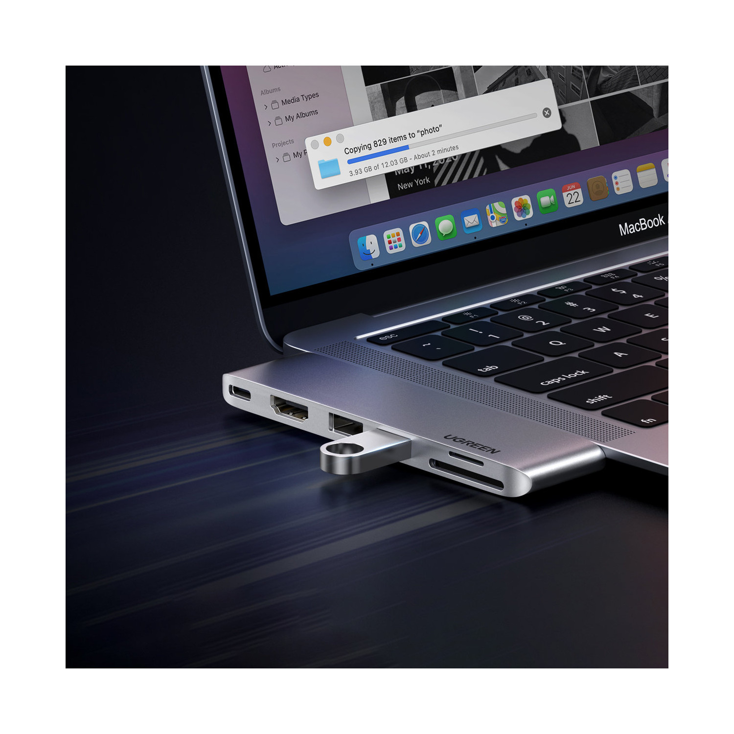 MacBook z dołączonym srebrnym hubem USB-C, pokazujący trwający transfer plików. Ekran wyświetla postęp kopiowania zdjęć.