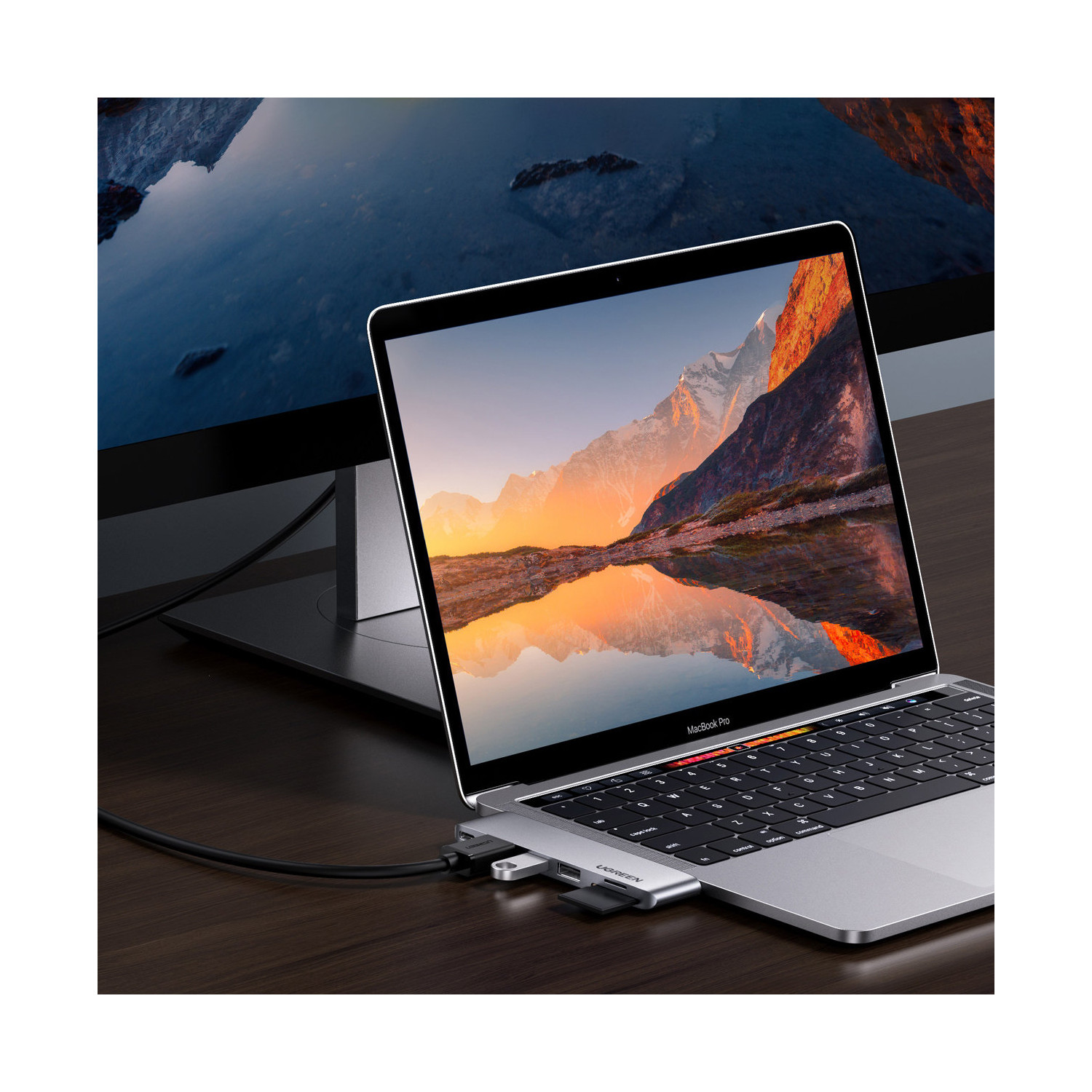 Laptop z górskim krajobrazem na ekranie, podłączony do huba USB i zewnętrznego monitora. Scena rozgrywa się na drewnianym stole.