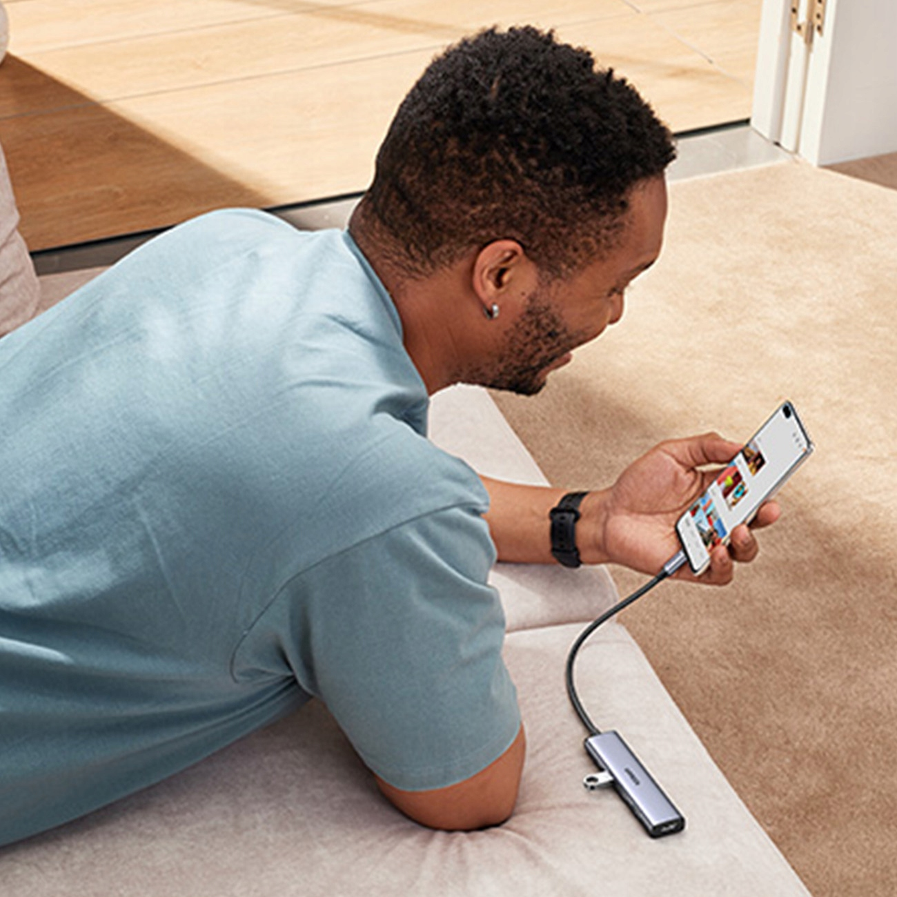 Mężczyzna leży na kanapie, patrząc na smartfon podłączony do koncentratora USB. Scena jest oświetlona naturalnym światłem.