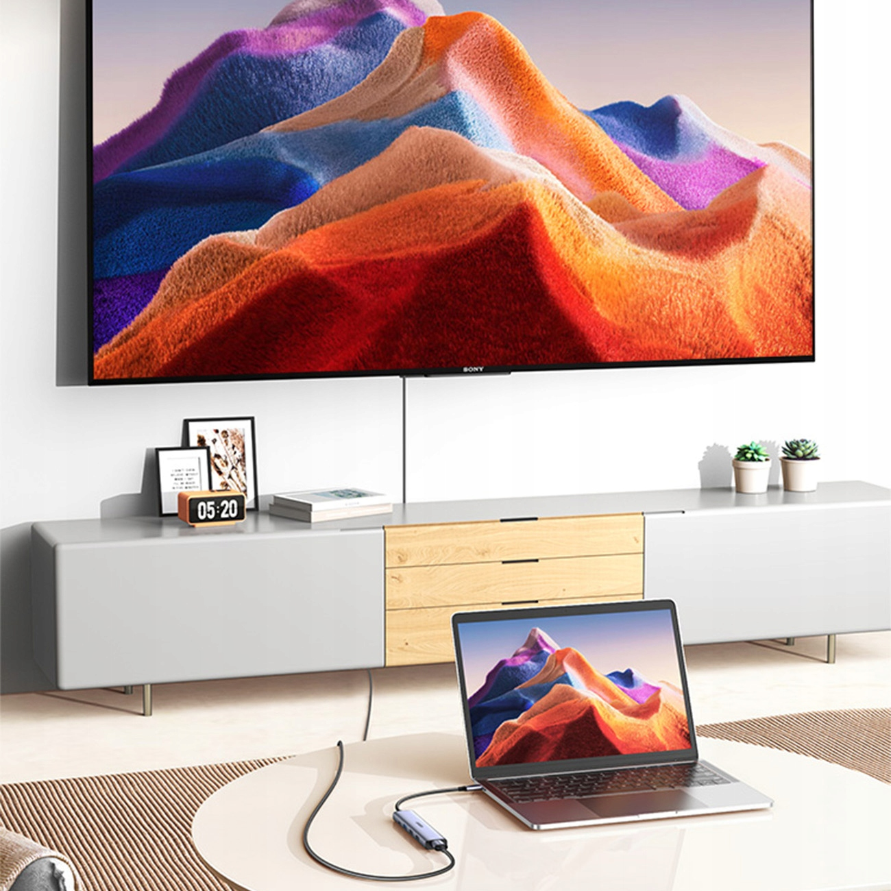 Laptop podłączony do telewizora wyświetlającego kolorową sztukę abstrakcyjną. Zegar i rośliny stoją na szafce pod telewizorem.