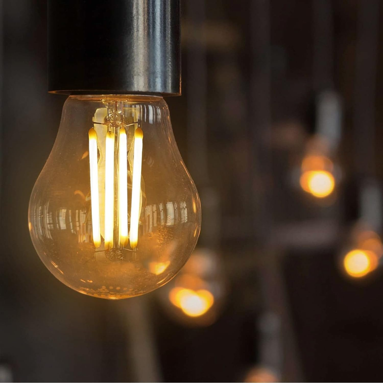Nahaufnahme einer beleuchteten Edison-Glühbirne mit schwarzem Anschluss, im Hintergrund verschwommene Lichter. Die Birne hat vertikale LED-Filamente.