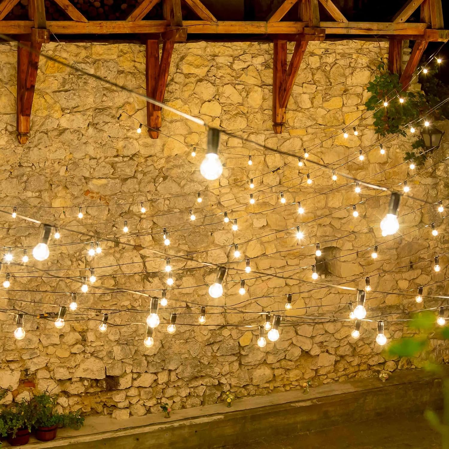 Lichterketten hängen vor einer Steinmauer. Holzbalken und Laub sind sichtbar. Die Lichter sind beleuchtet und erzeugen ein warmes Ambiente.