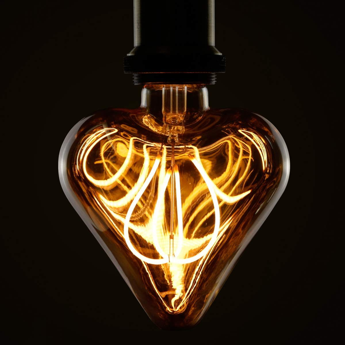 Żarówka w kształcie serca świeci ciepłym bursztynowym odcieniem. Jej włókna są skomplikowanie ułożone na ciemnym tle.