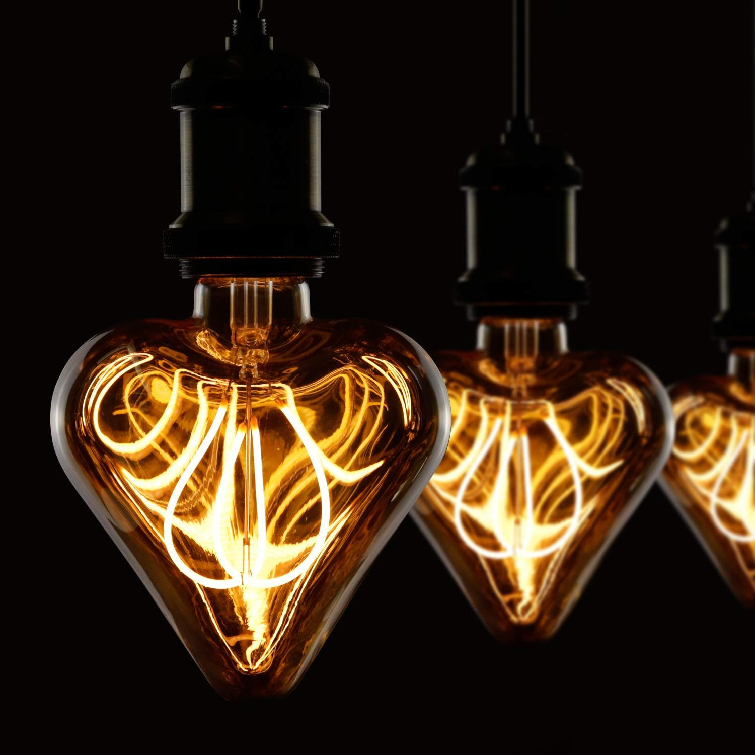 Trzy żarówki w kształcie serca z żarzącymi się włóknami wiszą na czarnym tle. Żarówki są w ciepłym bursztynowym kolorze.