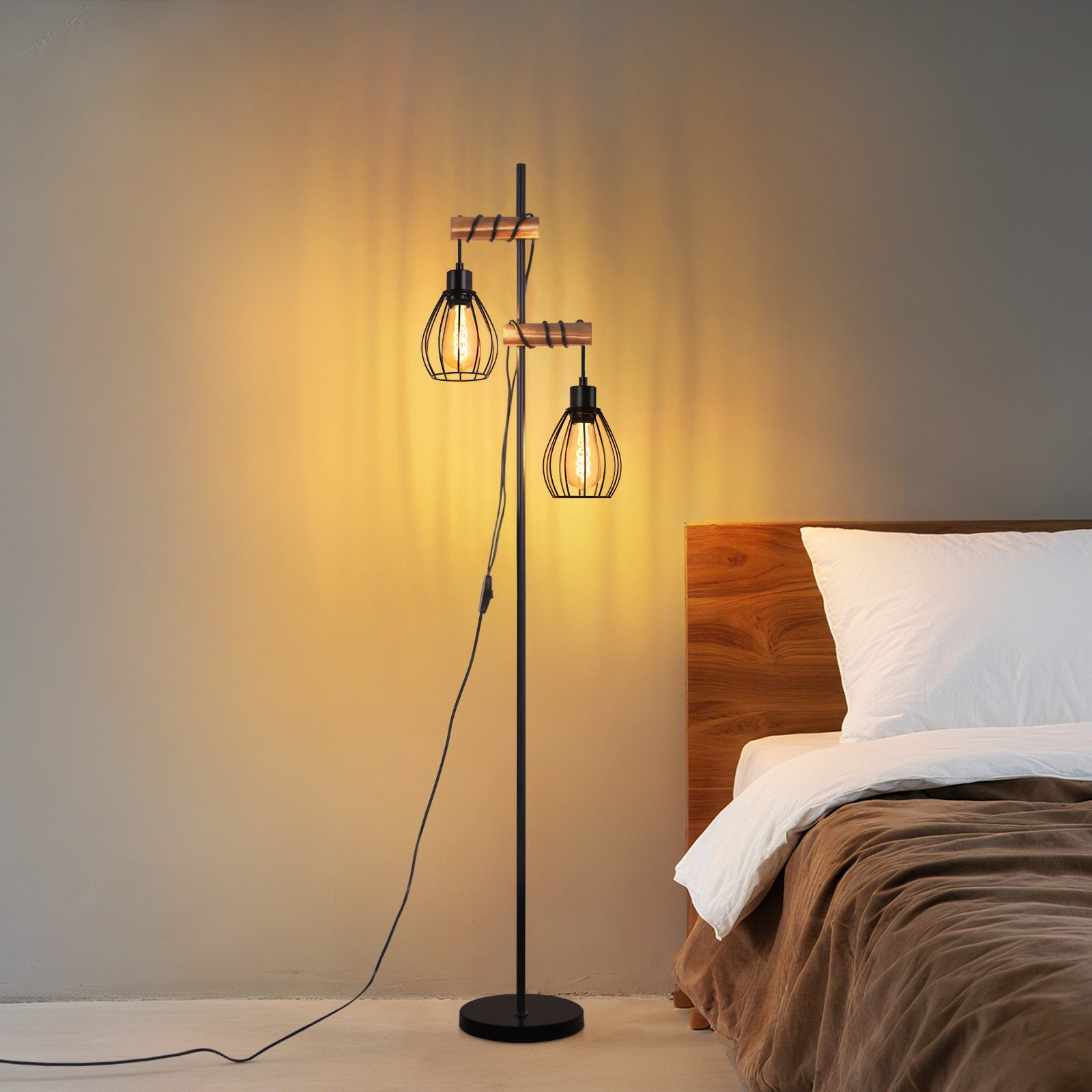 Lampa podłogowa z dwoma kloszami w kształcie klatki. Obok łóżka z białą poduszką i brązowym kocem. Ściana jest beżowa.