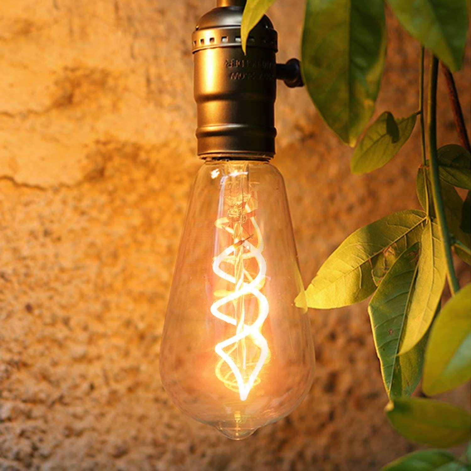 Świecąca żarówka Edisona wisi z zielonymi liśćmi po prawej stronie i teksturowaną brązową ścianą w tle. Żarówka ma spiralny żarnik.