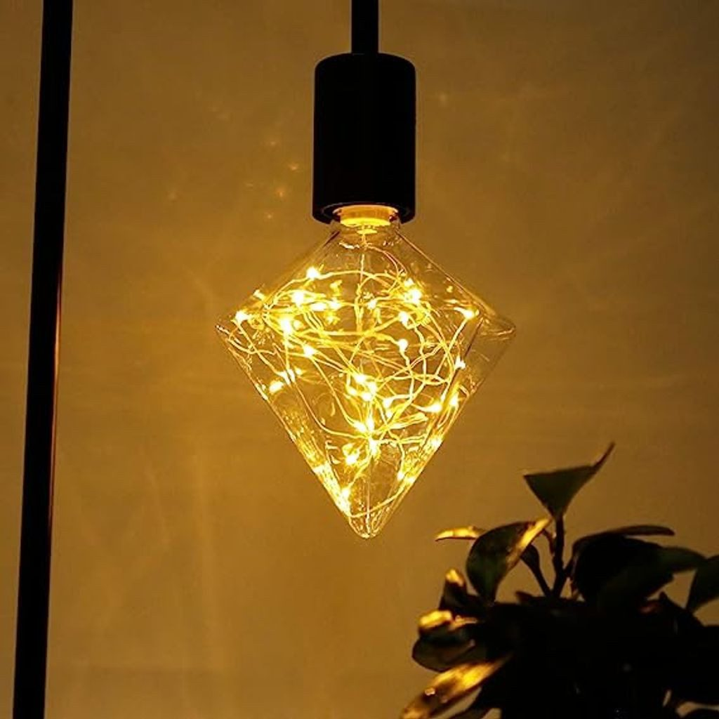 Żarówka w kształcie rombu z ciepłymi białymi światłami, czarne oprawy. Roślina w prawym dolnym rogu, żółte tło.