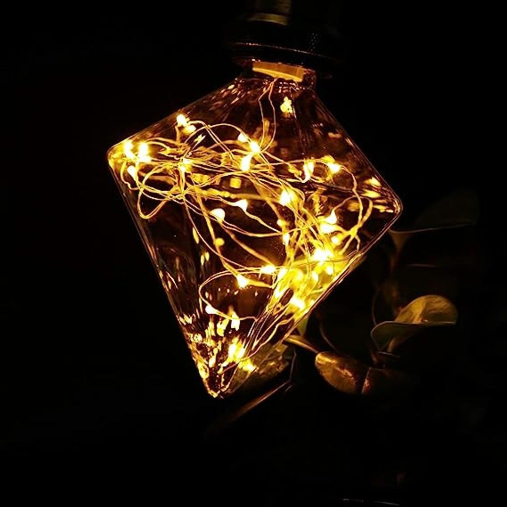 Ozdoba ze szkła w kształcie rombu wypełniona żółtymi lampkami, wisząca na czarnym tle z kilkoma zielonymi liśćmi.