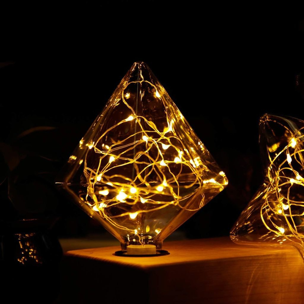 Dwie szklane lampy w kształcie rombu z ciepłymi światełkami w środku stoją na drewnianej powierzchni. Tło jest ciemne.
