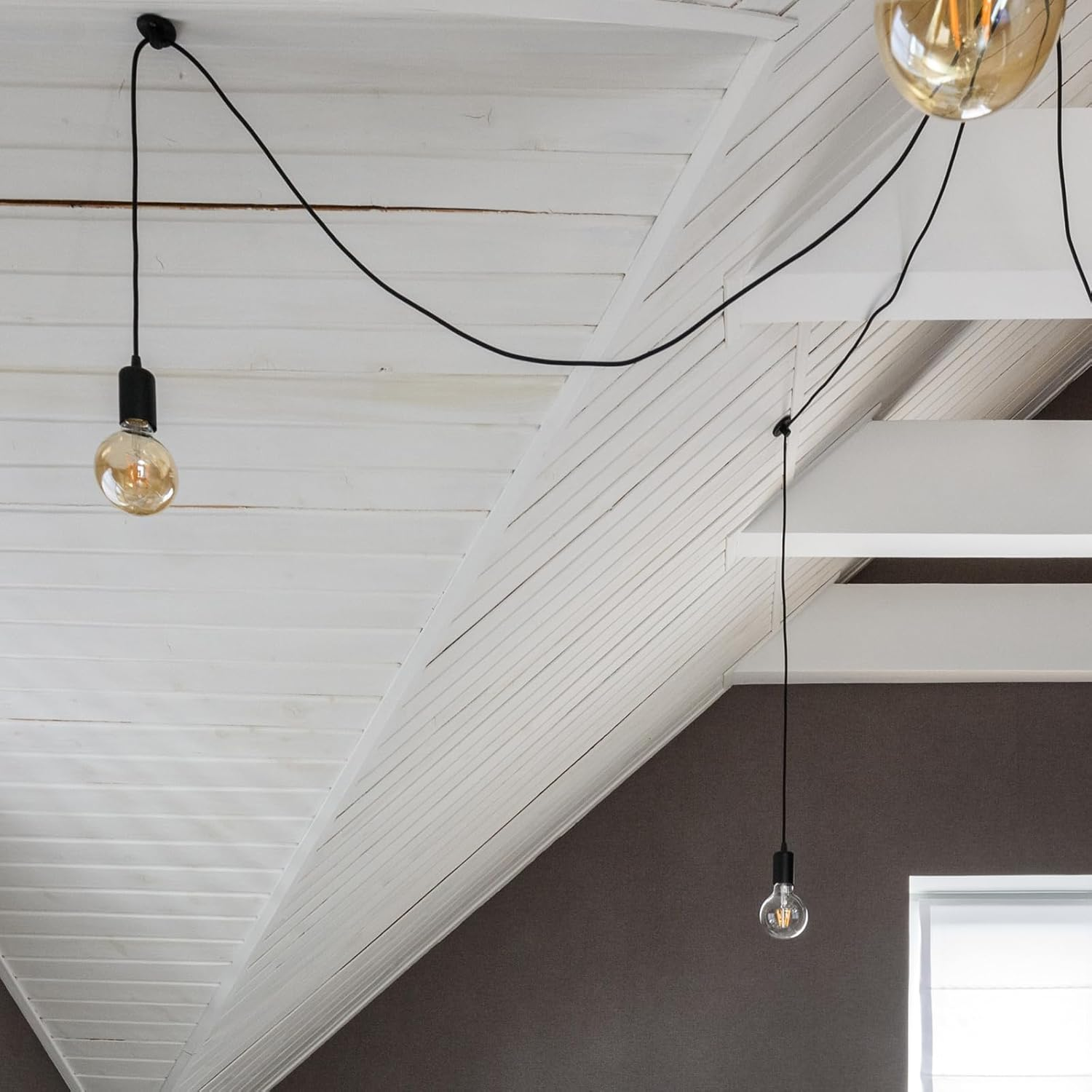 Biały sufit z listew z czarnymi przewodami i wiszącymi żarówkami. Jedna żarówka jest zapalona, ​​inne nie. Ciemnoszara ściana i rama okienna w tle.