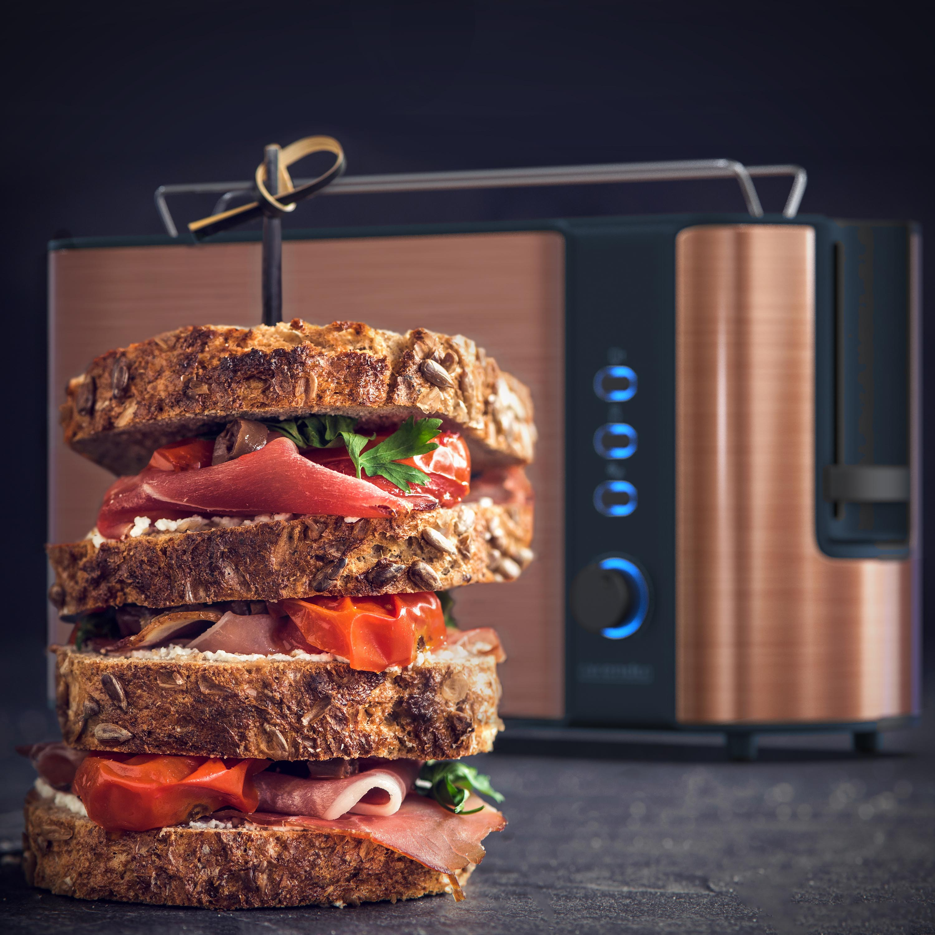 Geschichtetes Sandwich mit Tomaten, Schinken und Käse, gehalten von einem Holzspieß. Im Hintergrund ein Kupfertoaster.