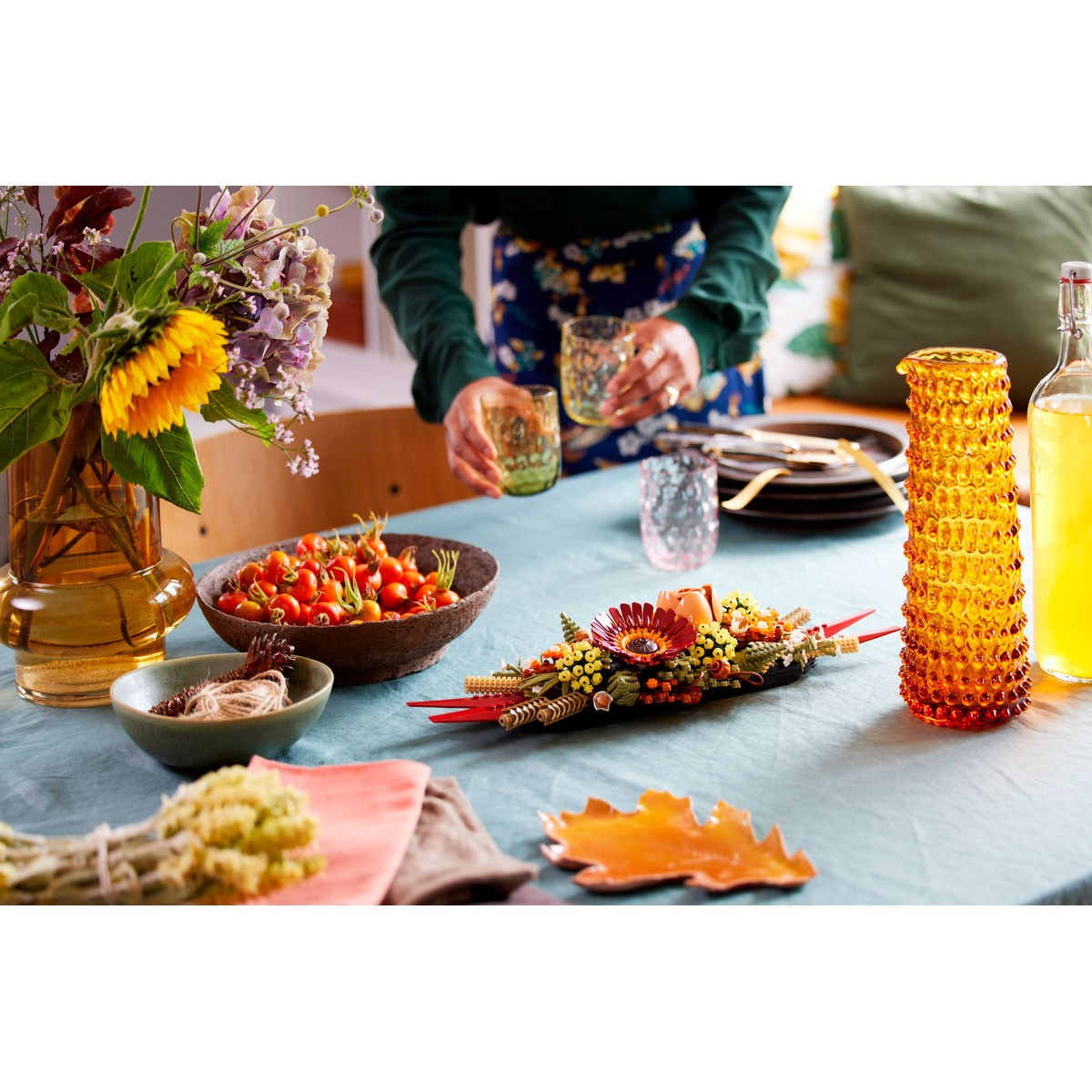 Une table garnie de fleurs, de fruits, de verres et d'une pièce maîtresse décorative. Une personne tient deux verres. Les couleurs incluent l'orange et le vert.