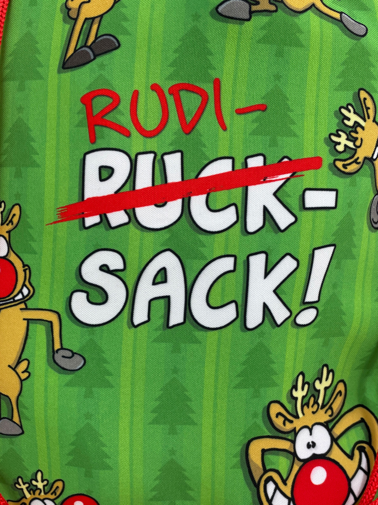 Grüner Rucksack mit Rentier-Muster. Text: 'Rudi- Ruck- Sack!' mit durchgestrichenem 'Ruck'. Rote Akzente.