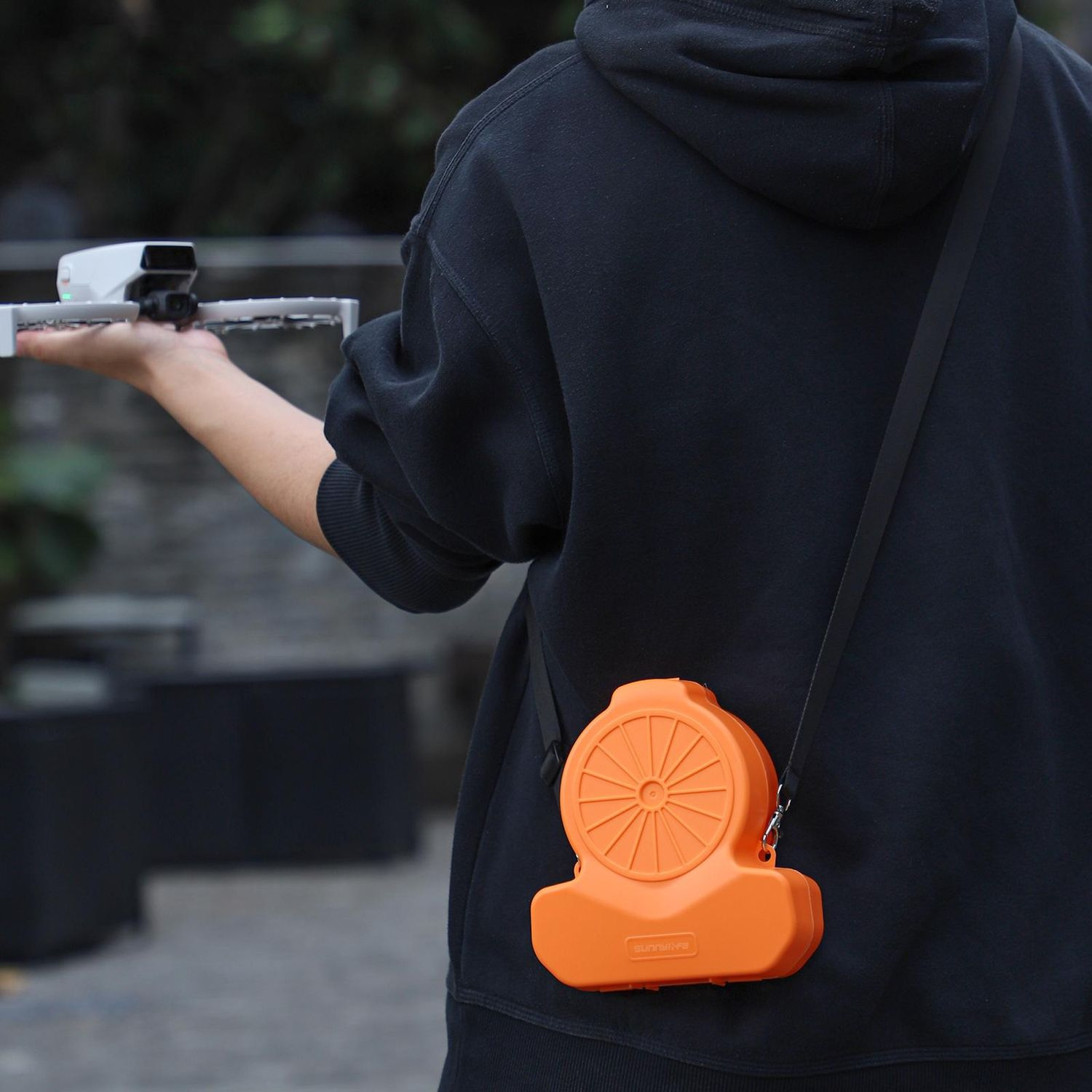 Person hält Drohne, trägt schwarzen Kapuzenpullover, mit orangefarbenem Gehäuse an einem Riemen. Hintergrund: verschwommene Außenszene.