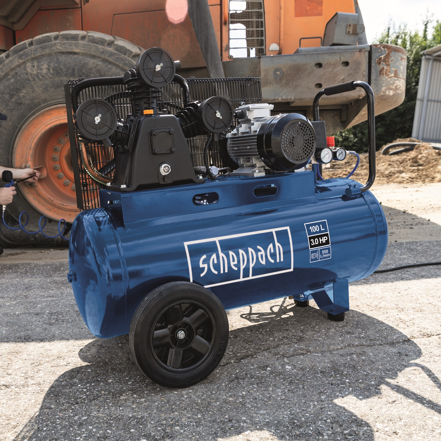 Een blauwe luchtcompressor met zwarte wielen. Een oranje zware machine op de achtergrond. Een persoon houdt een gereedschap vast.