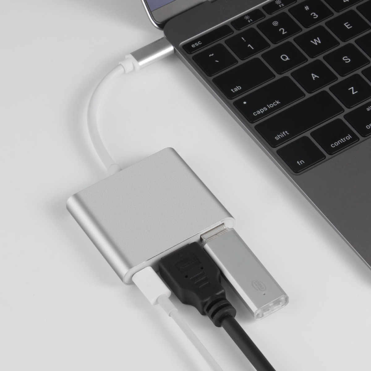 Laptop ist mit einem silbernen Hub mit Kabeln und einem USB-Laufwerk verbunden. Die Tastatur ist sichtbar. Weißer Hintergrund.