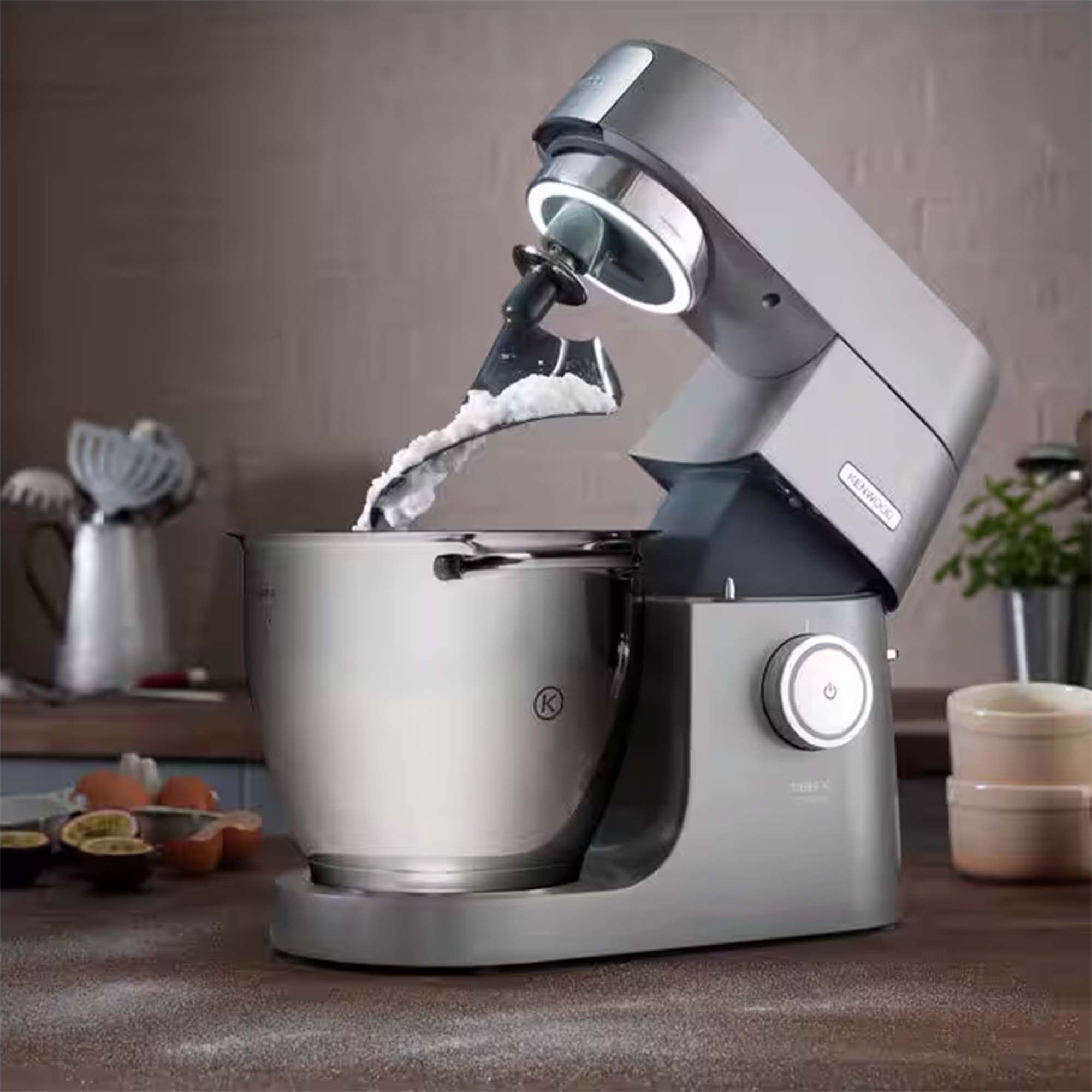 Un batteur KitchenAid argenté mélange de la pâte dans un bol. Œufs et fruits de la passion sur la table.