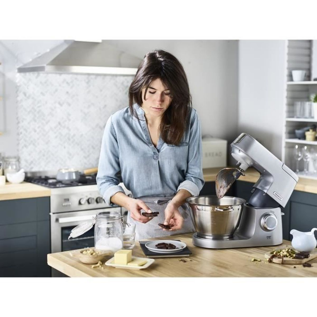Femme dans la cuisine avec un mixeur. Tient du chocolat. Ingrédients sur la table : noix, beurre, sucre. Arrière-plan : four, étagères, placards.