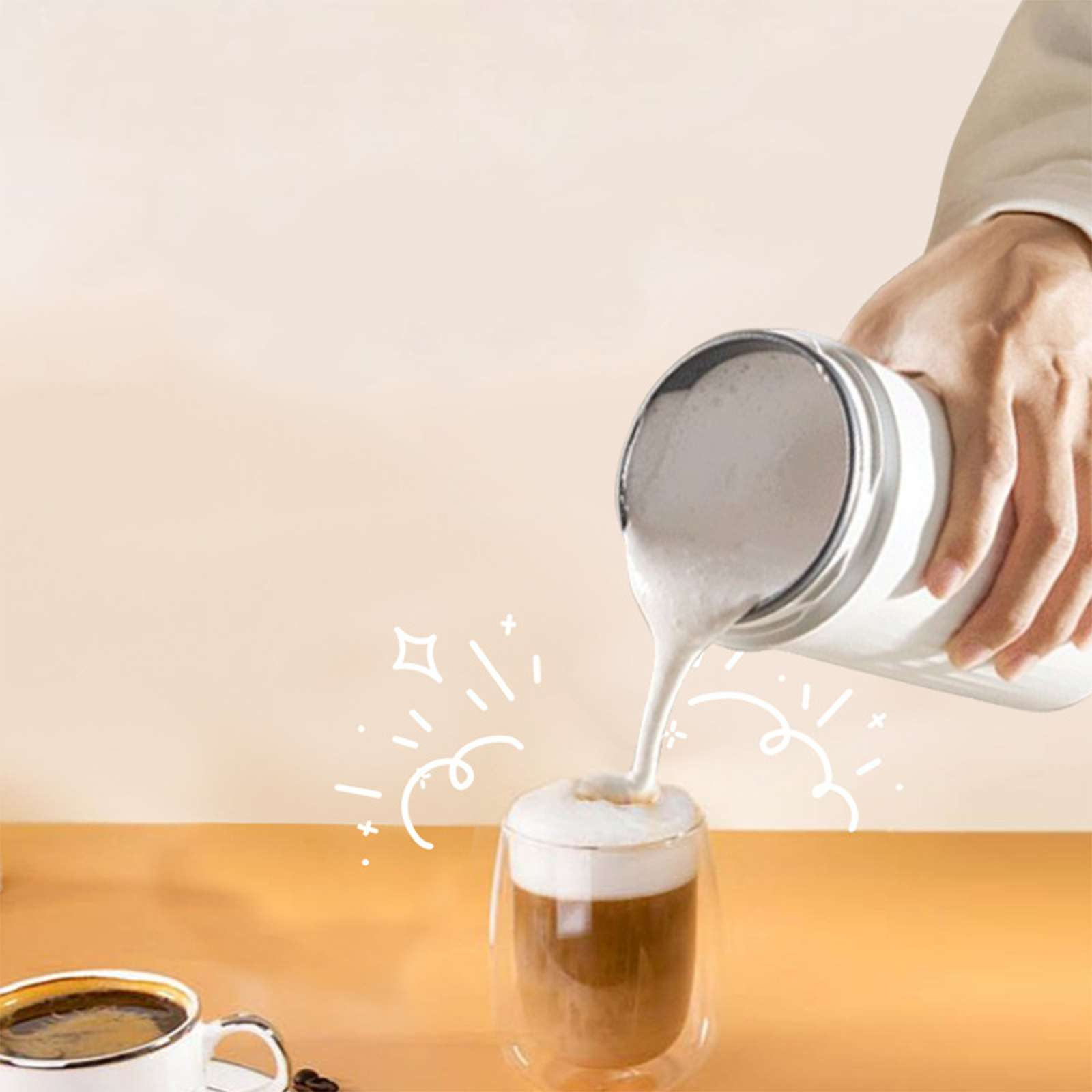 Eine Hand gießt Milchschaum in ein Kaffeeglas. Eine Tasse Kaffee steht links.