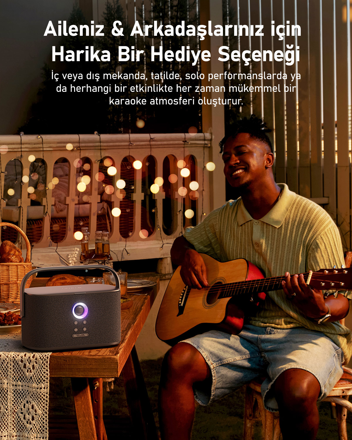Bir adam gitar çalıyor ve gülümsüyor. Bir hoparlör bir masada, arka planda ışıklar ve bir sepet var.