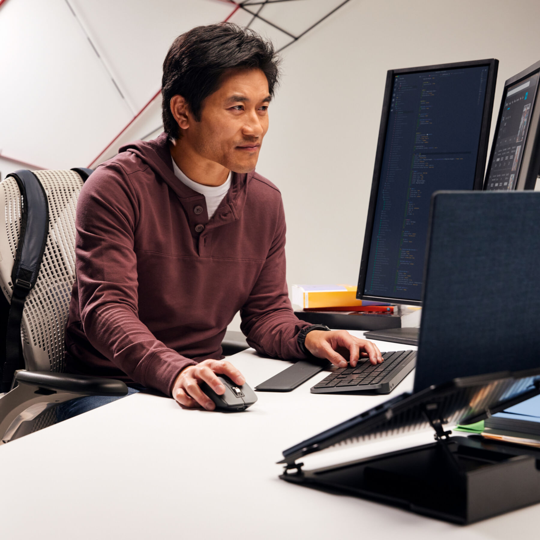 Mężczyzna pracuje przy biurku z dwoma monitorami, klawiaturą i myszą. Ma na sobie bordową bluzę z kapturem. Tło to biała ściana.