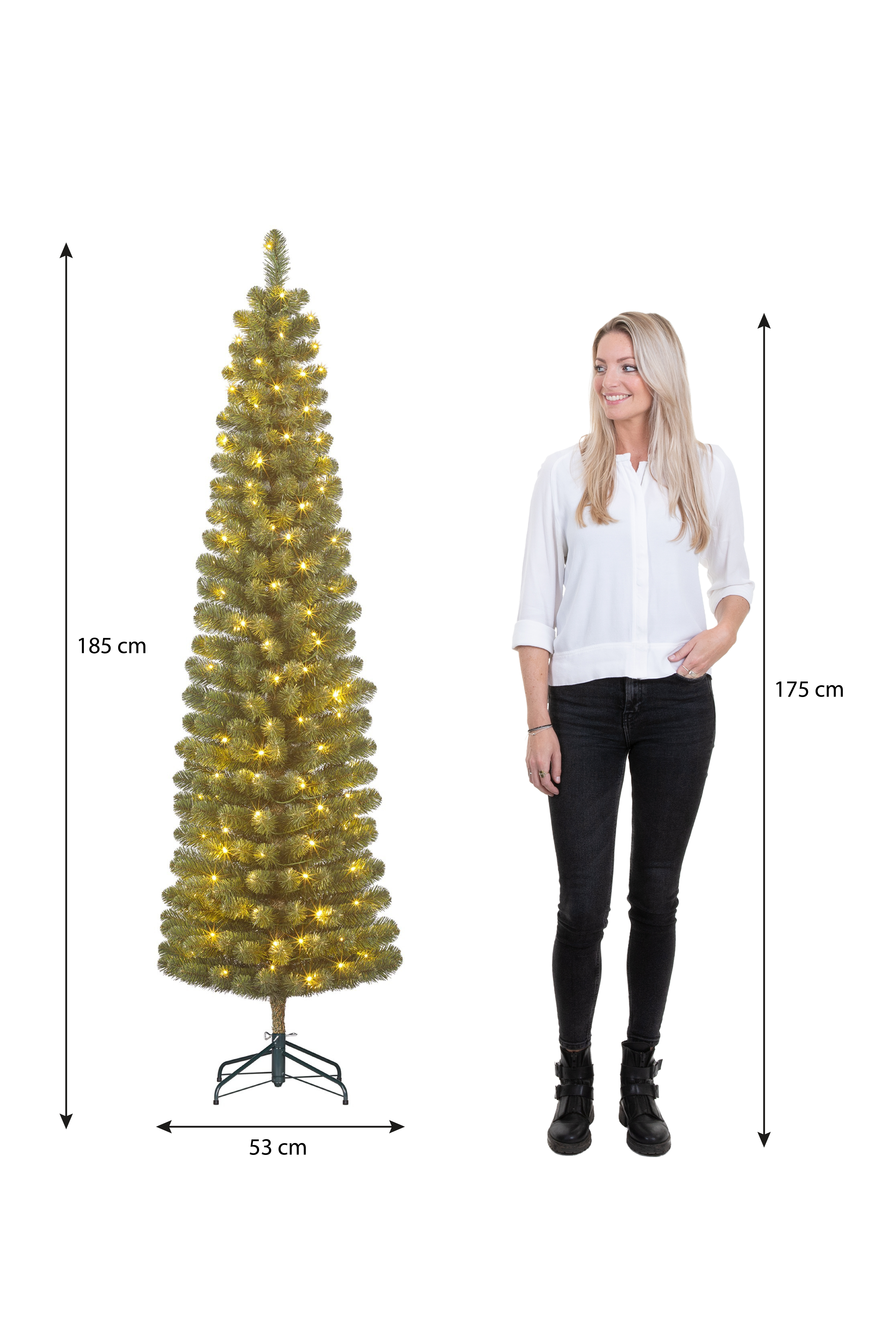 Une femme se tient à côté d'un sapin de Noël. L'arbre est éclairé et la femme porte une chemise blanche et un pantalon noir.