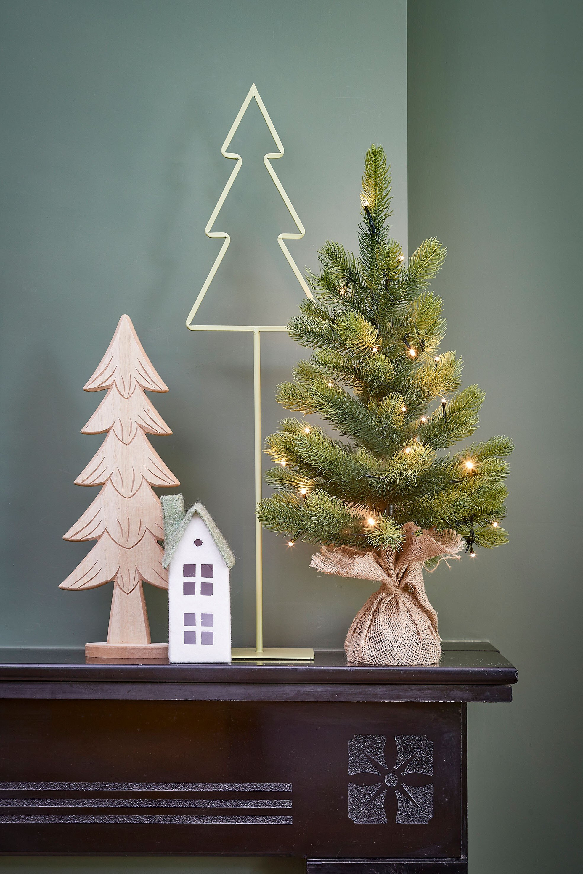 Décorations de Noël sur une surface marron foncé. Un petit arbre avec des lumières, un arbre en bois, une maison et un arbre en métal sont présents.