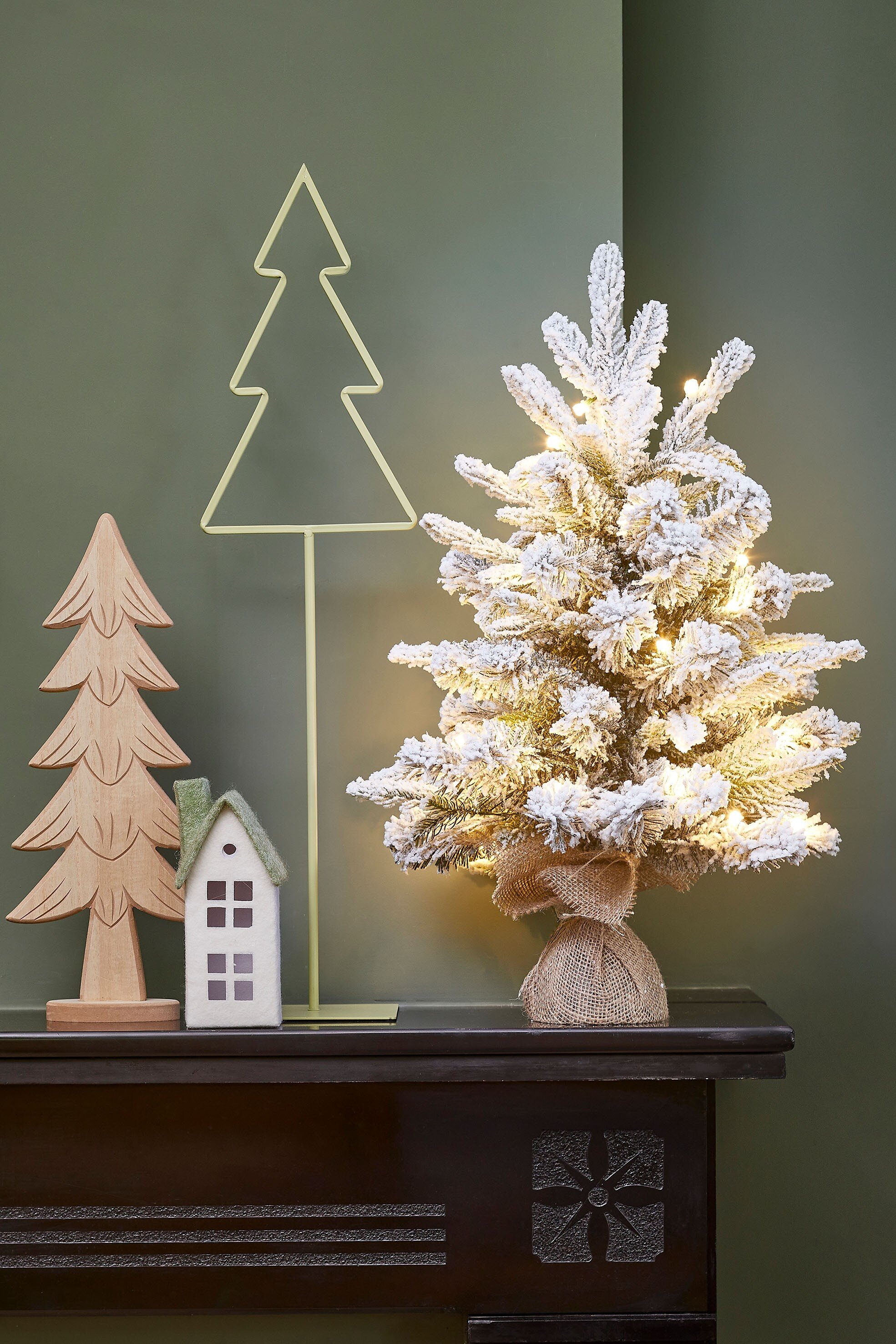 Une scène de Noël décorée. Comprend un arbre enneigé, un arbre en bois, une maison et un contour d'arbre, sur une étagère sombre.