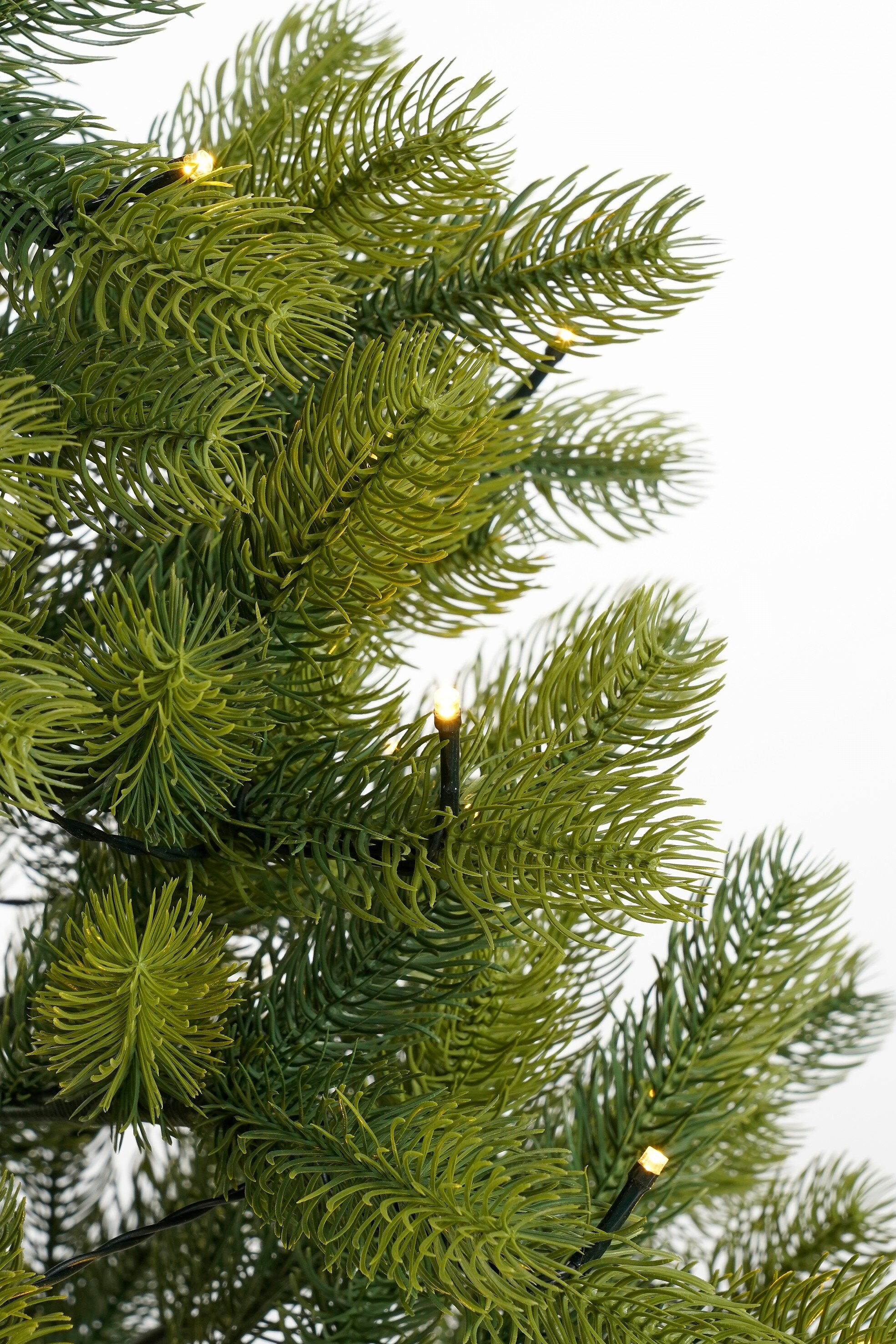 Gros plan sur un sapin de Noël artificiel vert. Quelques lumières sont allumées. Fond blanc.