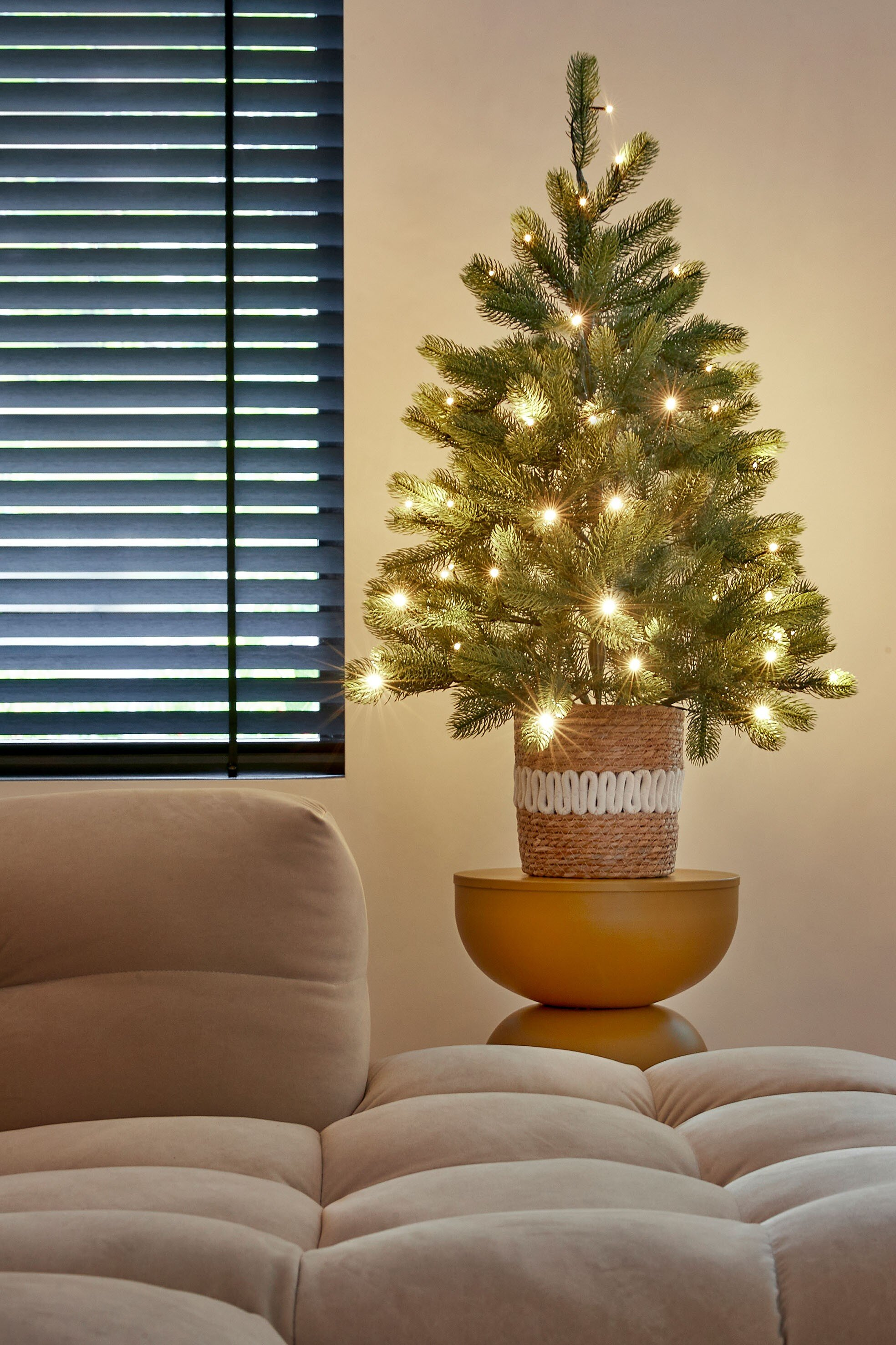 Un petit sapin de Noël avec des lumières est posé sur une table jaune. Un canapé beige est au premier plan, et une fenêtre à gauche.