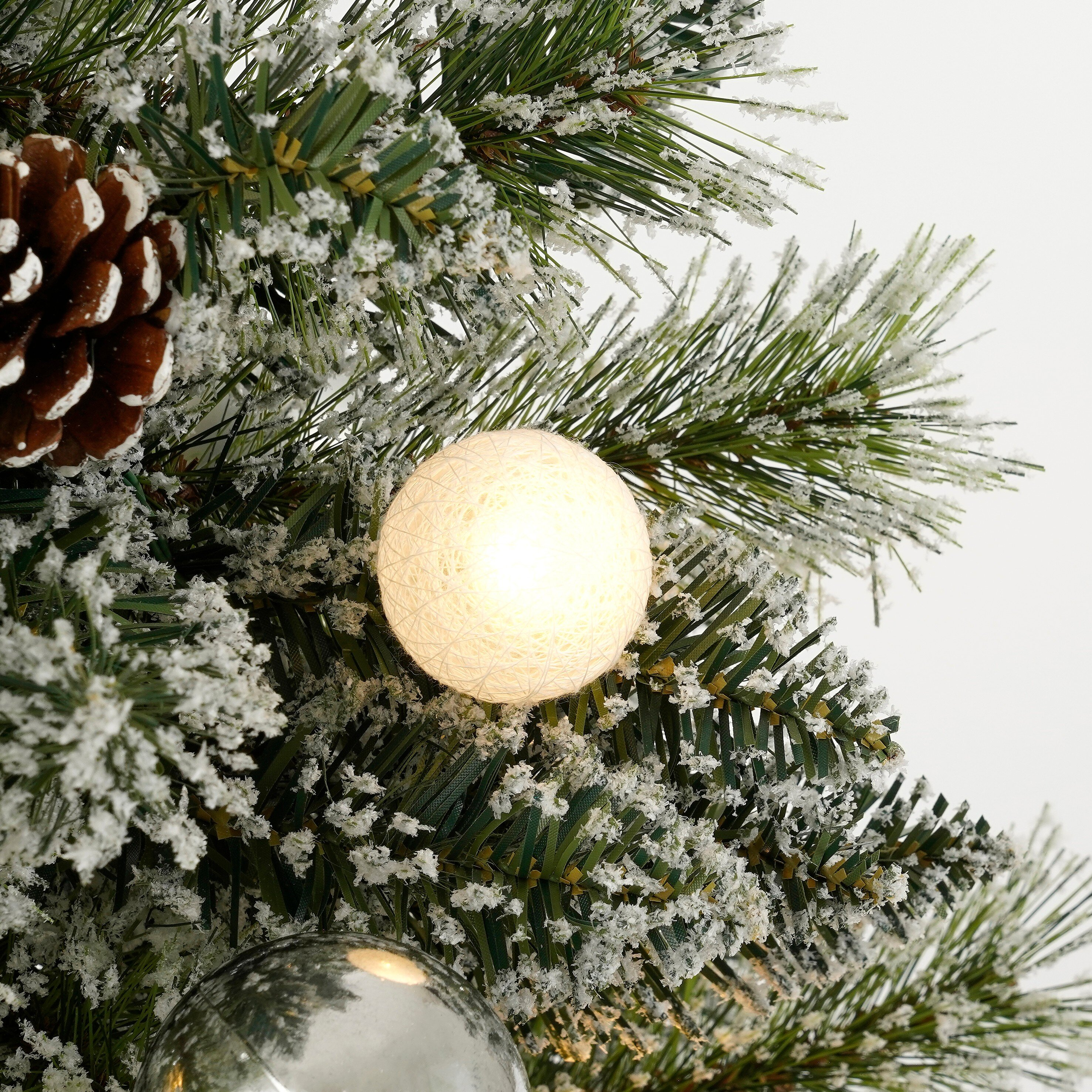 Close-up van een kerstboom. Hij heeft een dennenappel, een gloeiende bal en een zilveren bal. De boom is bedekt met sneeuw.