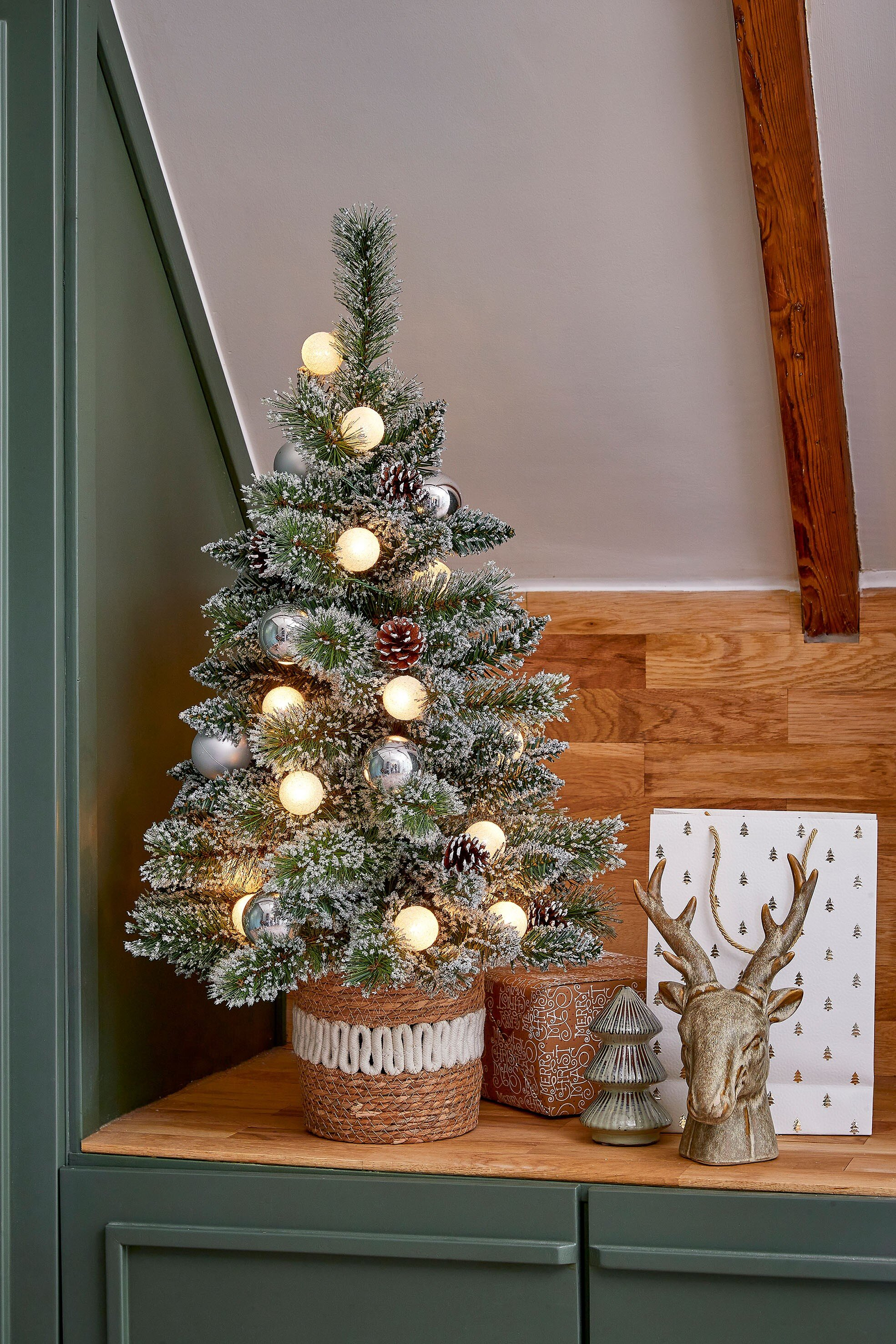 Een versierde kerstboom met lichtjes, ornamenten en dennenappels staat op een houten oppervlak met een hertenkopstandbeeld.