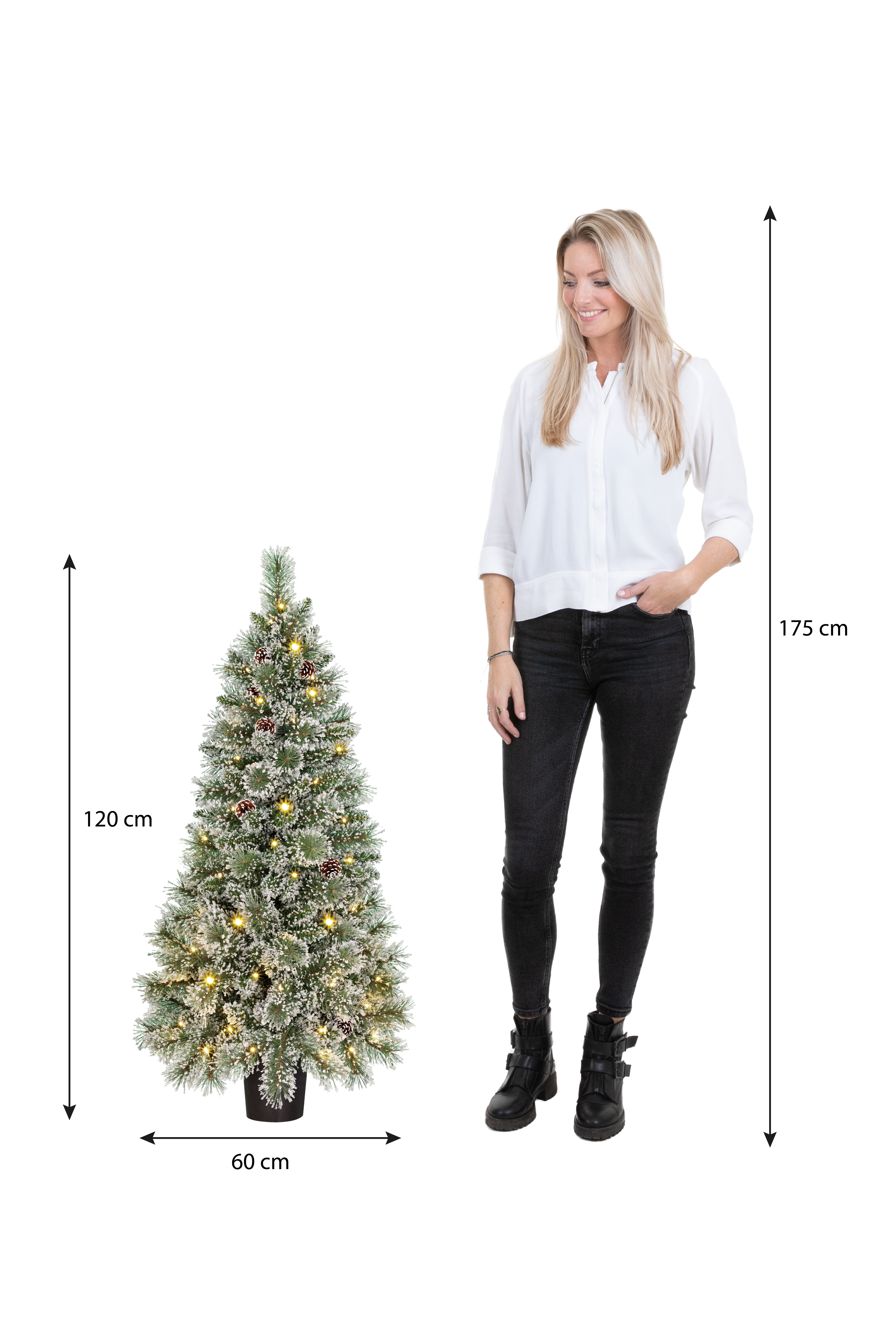 Een vrouw staat bij een kerstboom. De boom is versierd, de vrouw draagt een wit overhemd en een zwarte jeans.