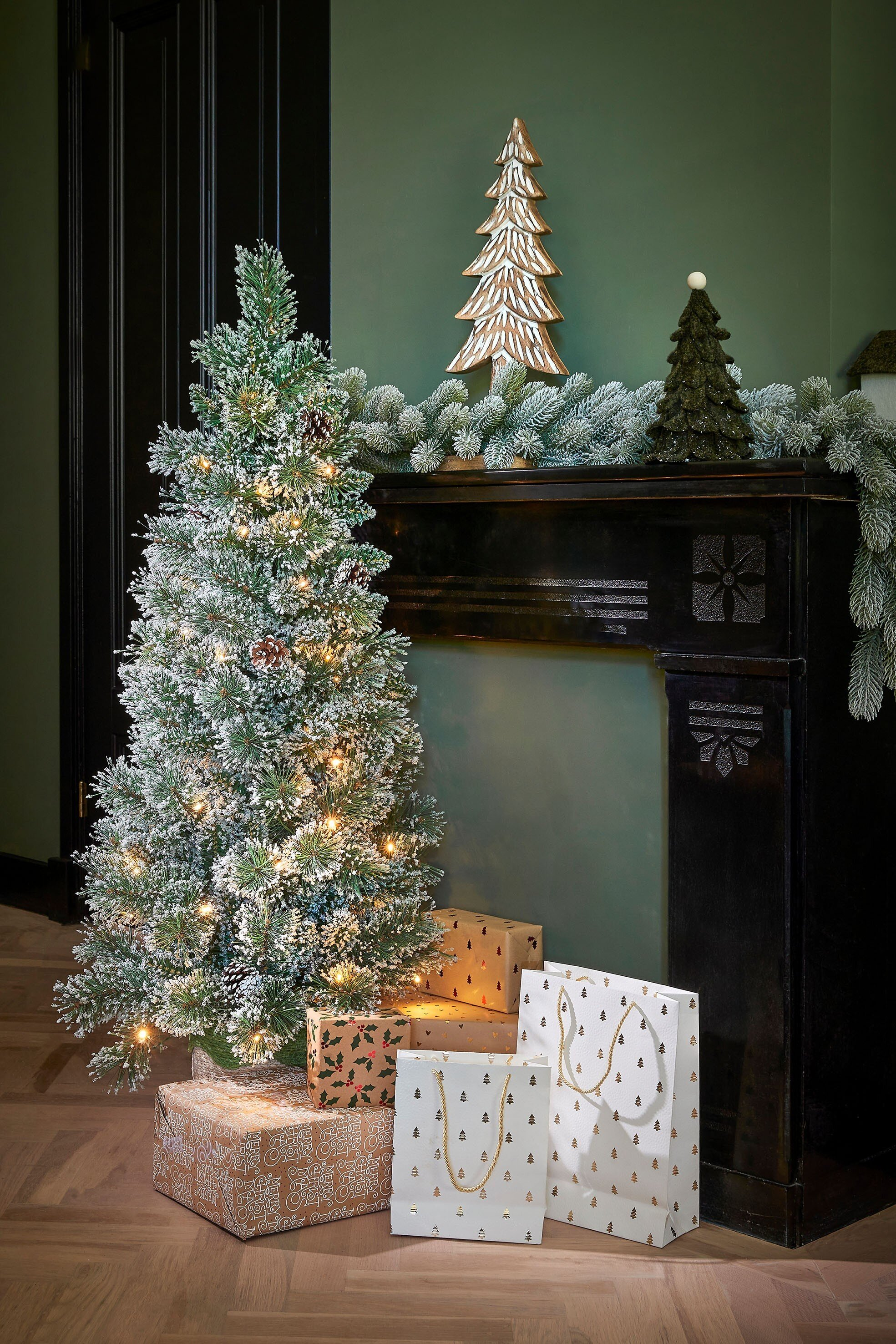 Een versierde kerstboom met cadeaus, papieren tassen en feestelijke decoraties bij een open haard.