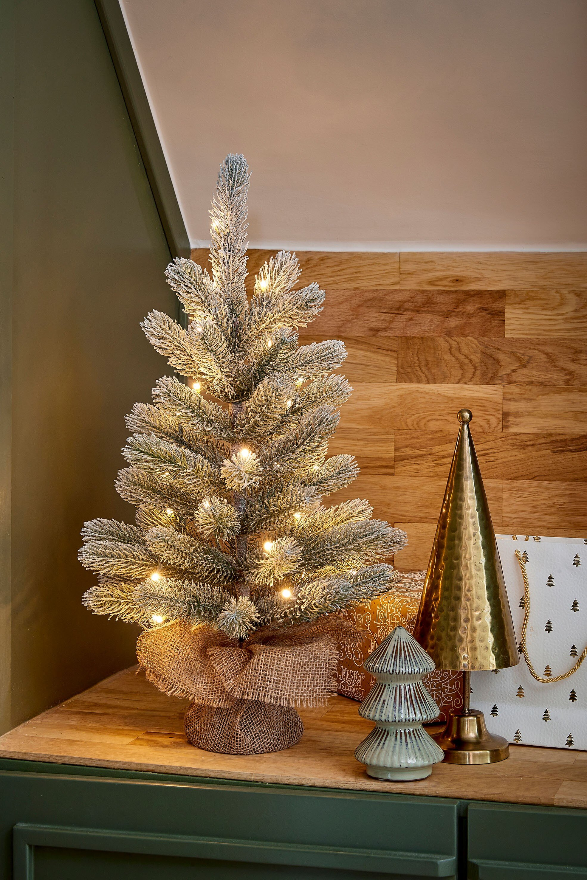 Un petit sapin de Noël avec des lumières. À côté, une décoration dorée en forme d'arbre et un sac cadeau.
