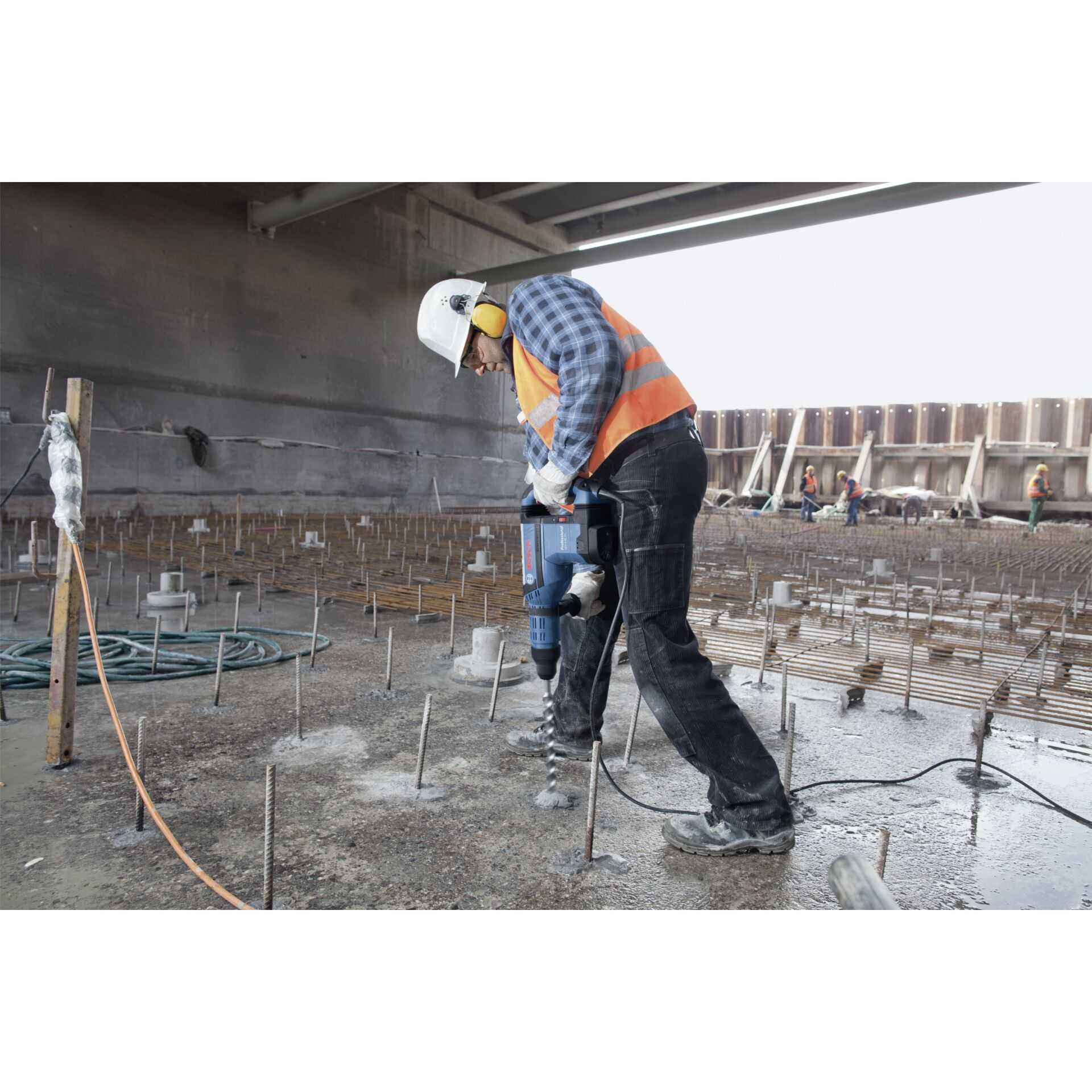 Ein Bauarbeiter bohrt mit Schutzkleidung und einem großen Bohrer in Beton. Bewehrungsstahl und andere Arbeiter sind sichtbar.