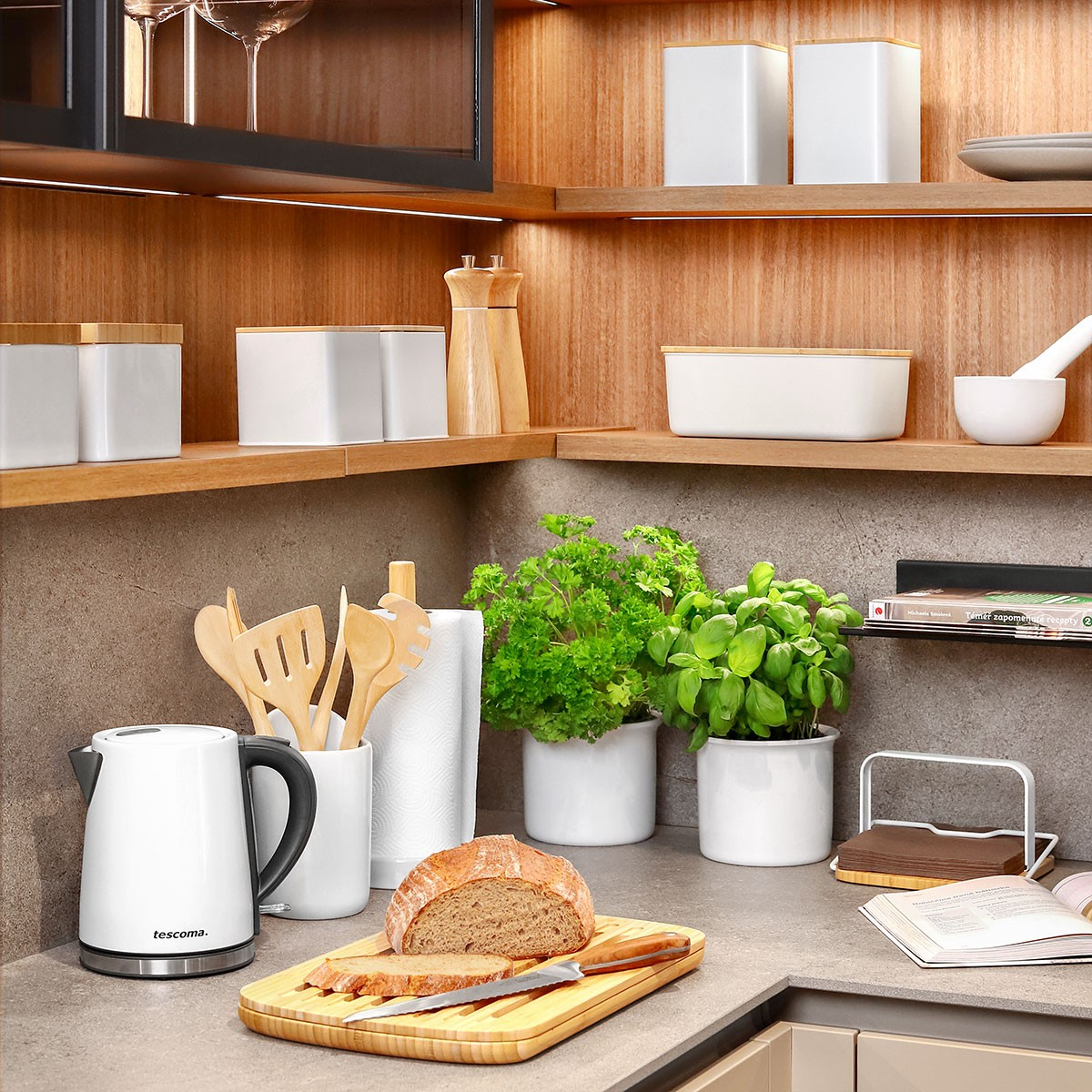 Eine Küchenzeile mit Wasserkocher, Brot, Pflanzen und Utensilien. Regale beherbergen Behälter und Gewürze, mit einem Brotbrett im Vordergrund.