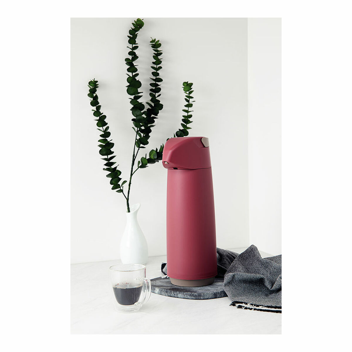 Eine maroonfarbene Thermoskanne, ein klares Kaffeeglas, Eukalyptus in einer weißen Vase und ein graues Tuch auf weißer Oberfläche.