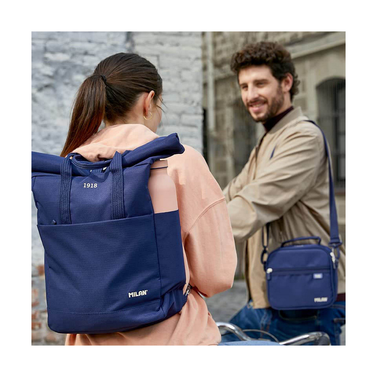 Eine Frau mit blauem Rucksack und ein Mann mit Umhängetasche lächeln. Beide Taschen sind mit MILAN versehen. Der Hintergrund ist eine Straße.