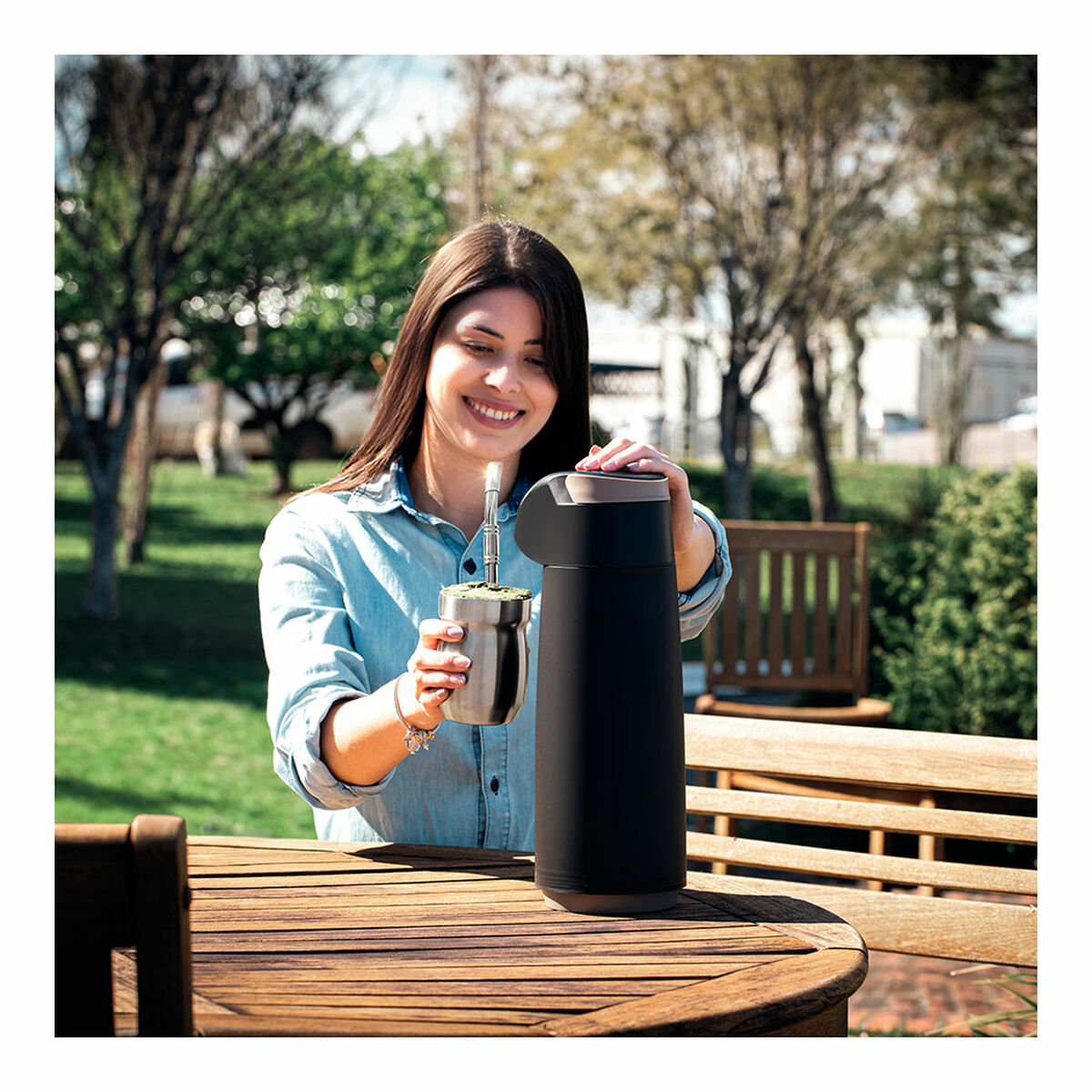 Frau in blauem Hemd lächelt, hält Mate-Becher und schwarze Thermoskanne auf einem Holztisch. Bäume und Büsche im Hintergrund.