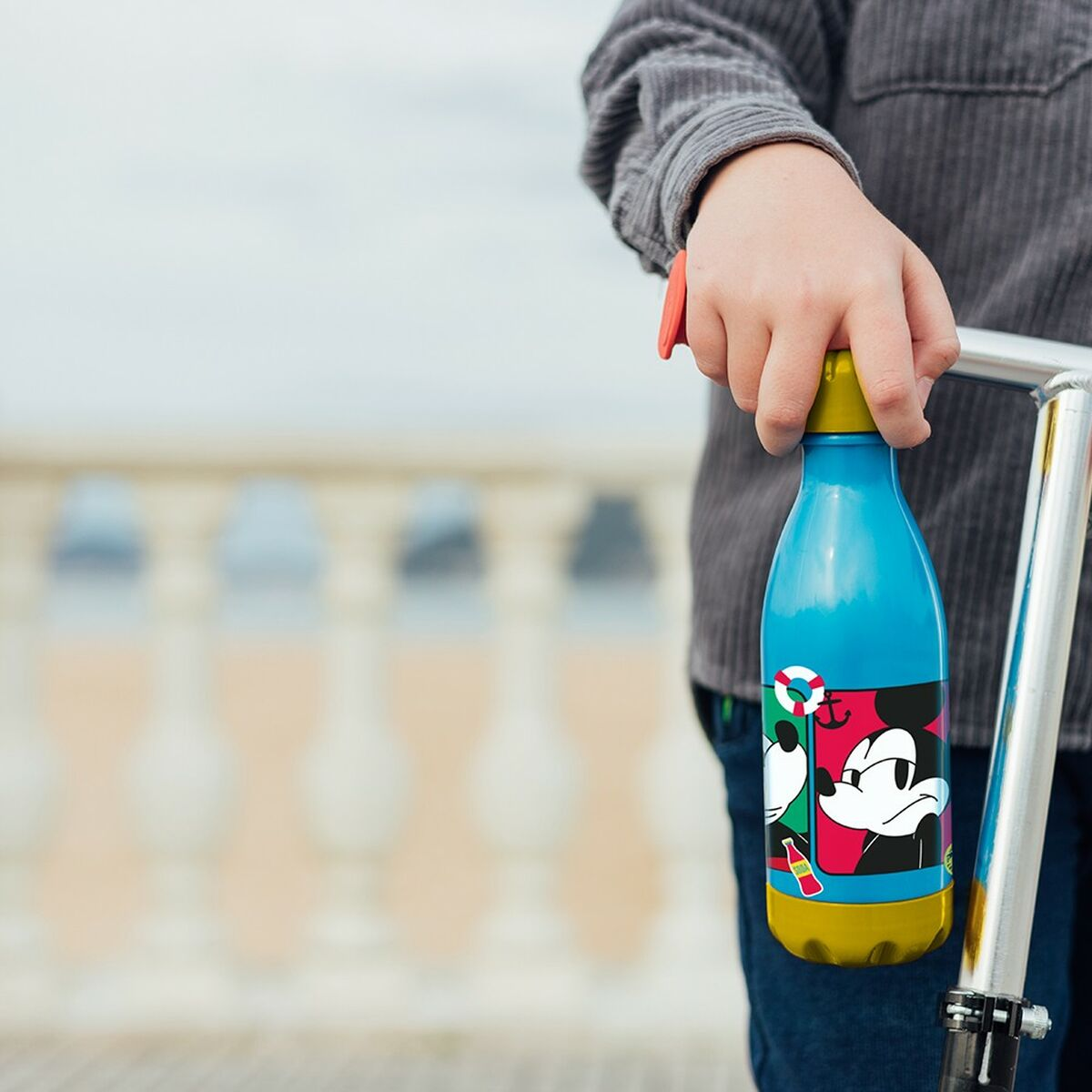 Ein Kind hält eine blaue Wasserflasche mit Mickey Mouse-Design. Die Flasche hat gelbe und grüne Akzente. Der Hintergrund ist unscharf.