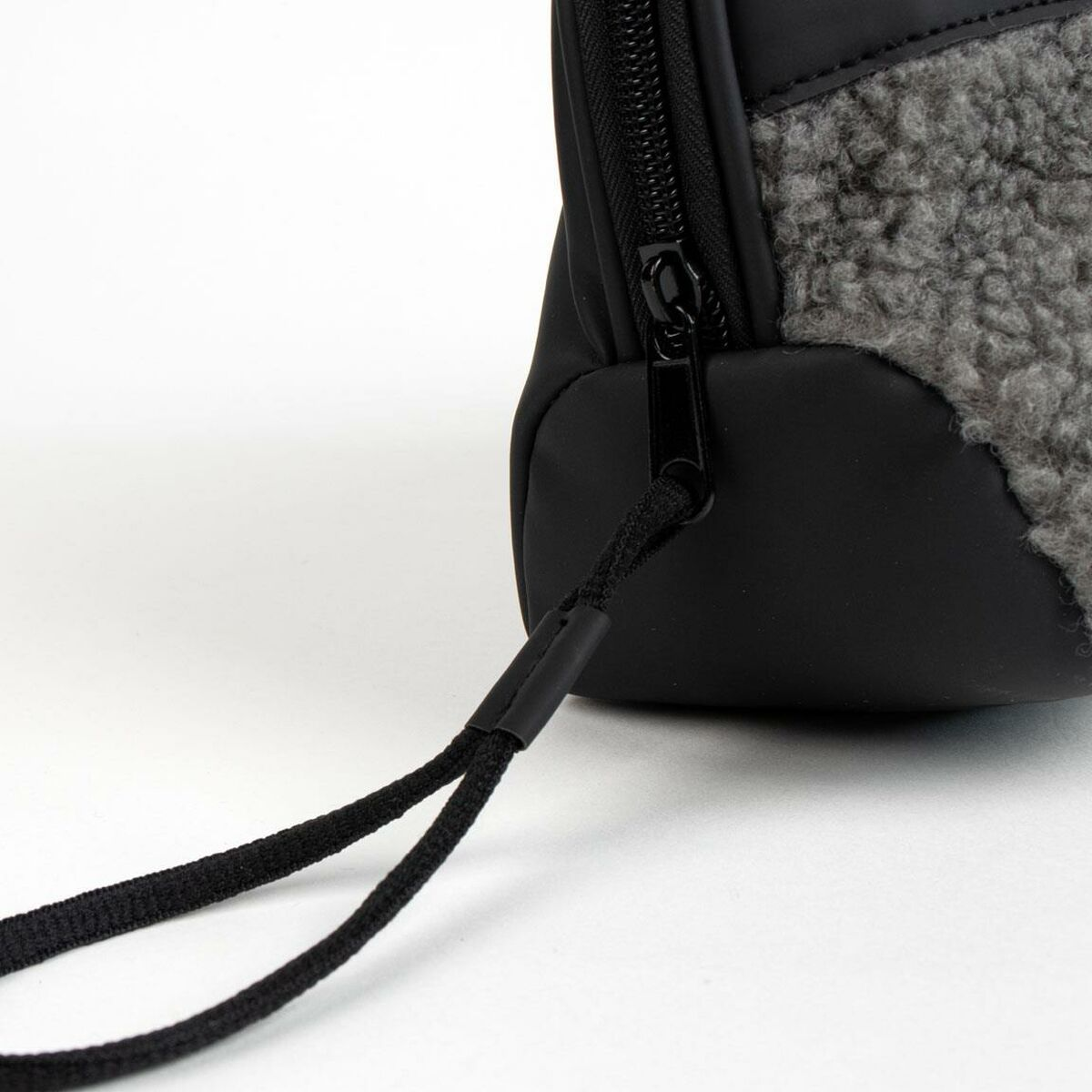 Nahaufnahme einer schwarzen Tasche mit grauer, pelziger Textur an der Seite und einem schwarzen Reißverschluss. Ein schwarzer Riemen geht von der Tasche aus.