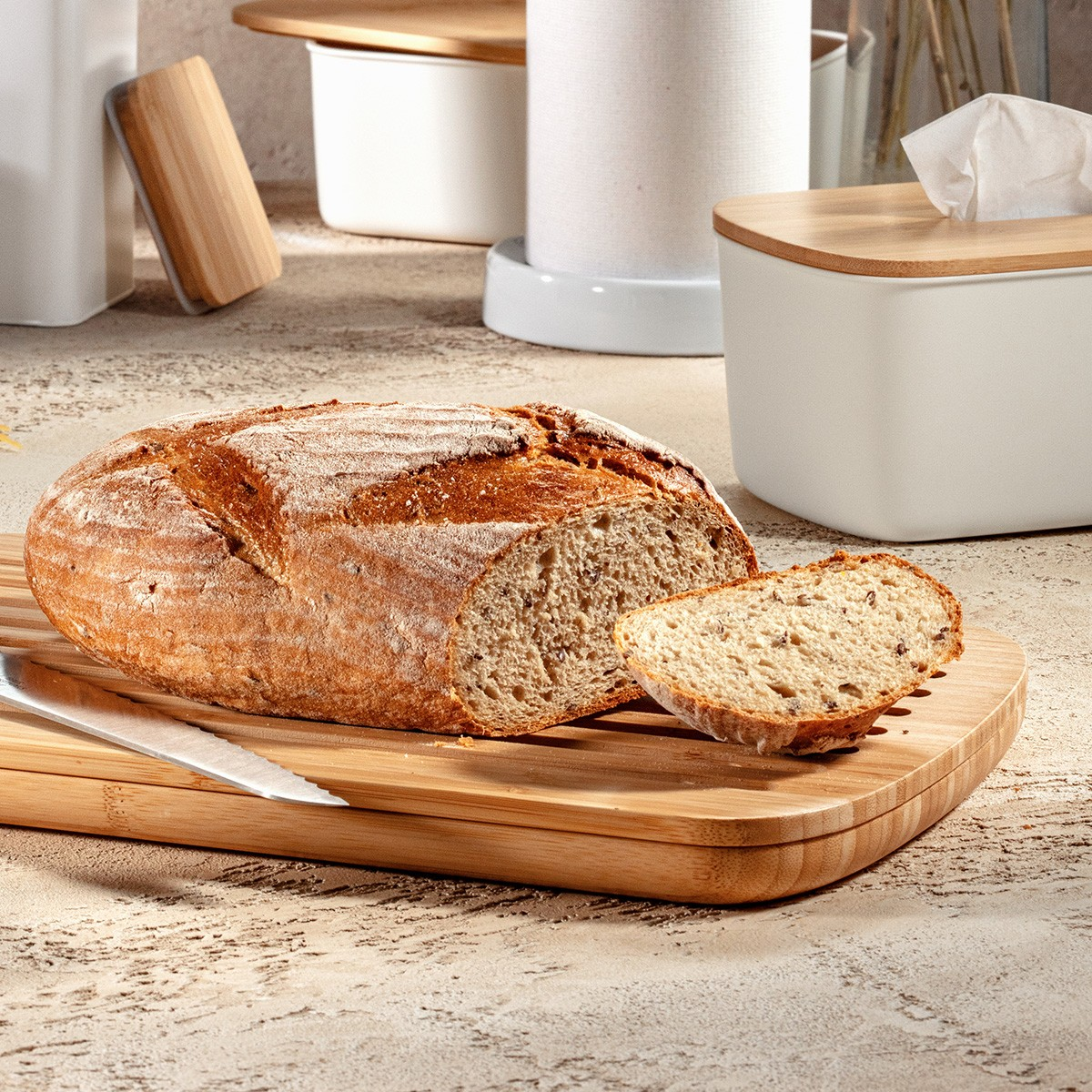Ein Brot, teilweise geschnitten, auf einem Holzbrett. Ein Messer liegt auf dem Brett. Weiße Behälter sind im Hintergrund.
