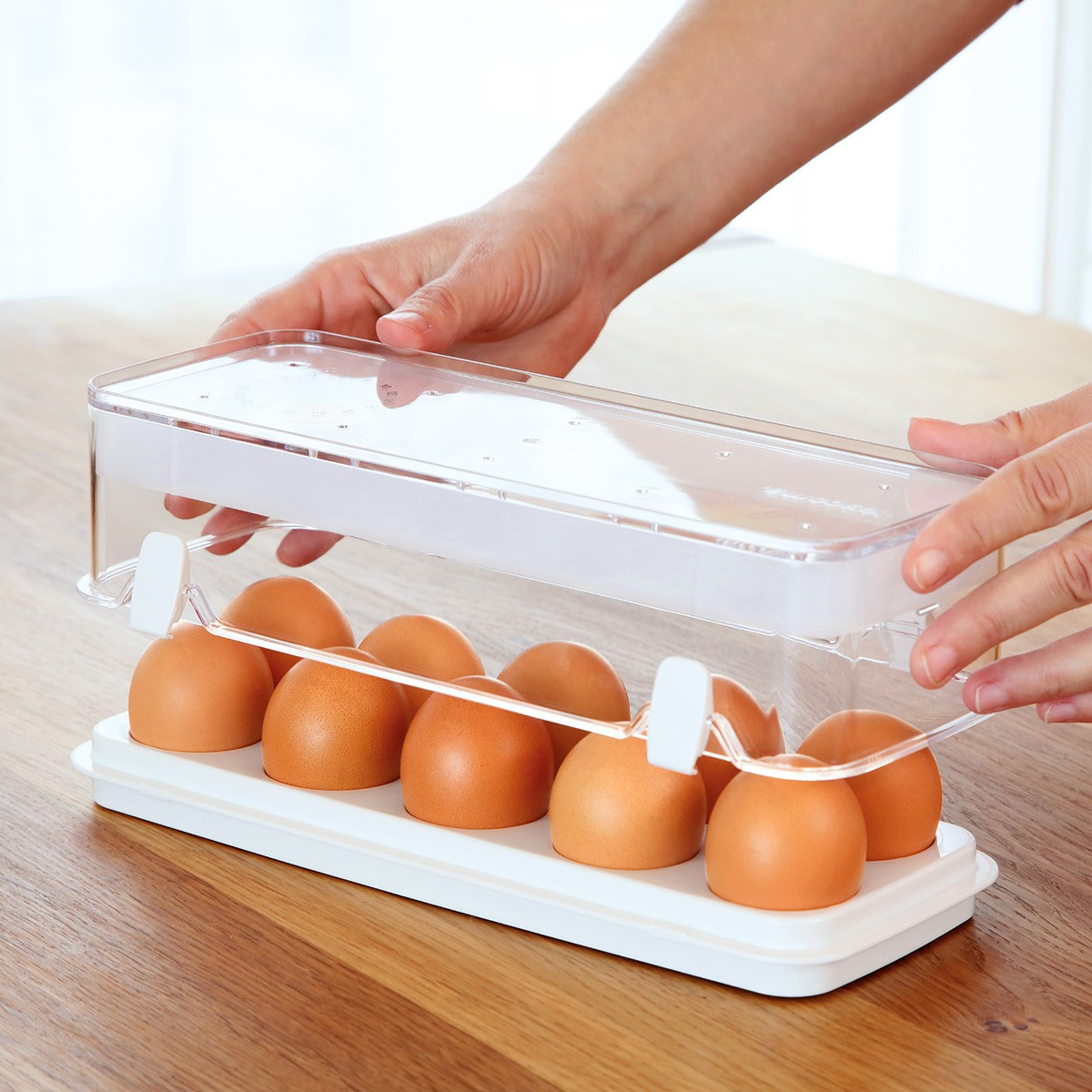 Una persona posiziona un portauova trasparente di plastica su una superficie di legno. Il supporto contiene uova marroni e ha base e coperchio bianchi.