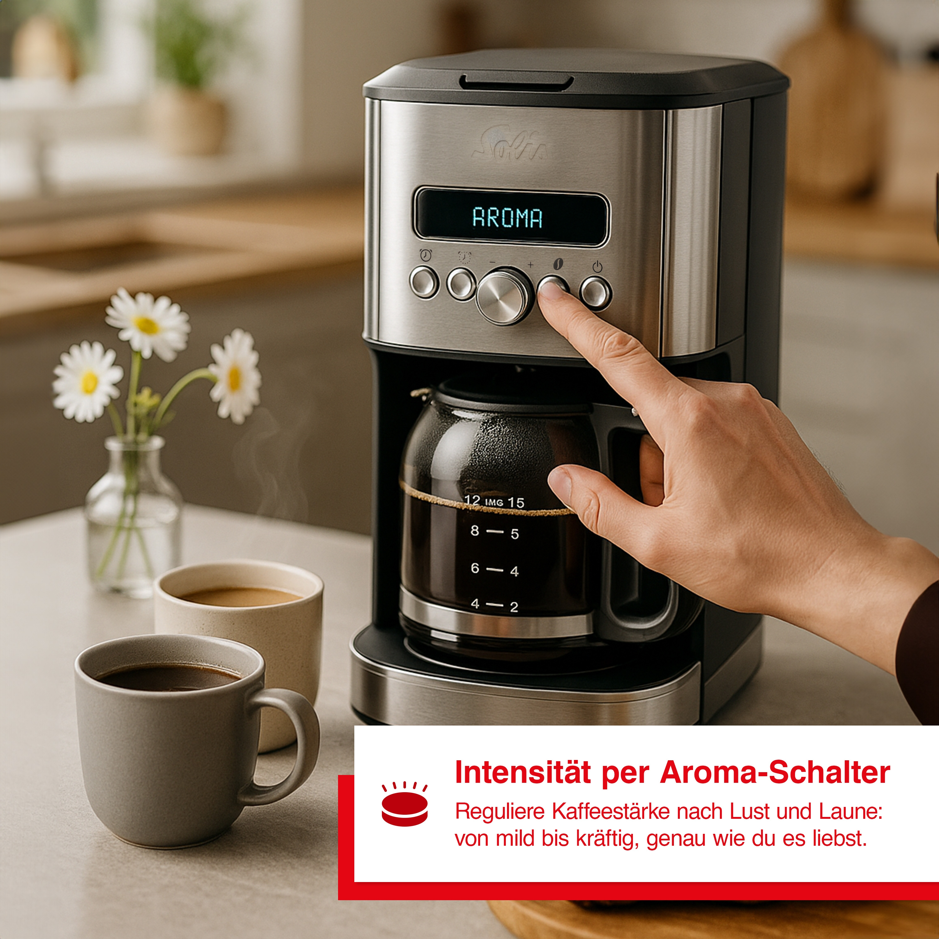 Kaffeemaschine mit Hand, die einen Knopf drückt. Zwei Tassen und Blumen stehen auf dem Tisch.